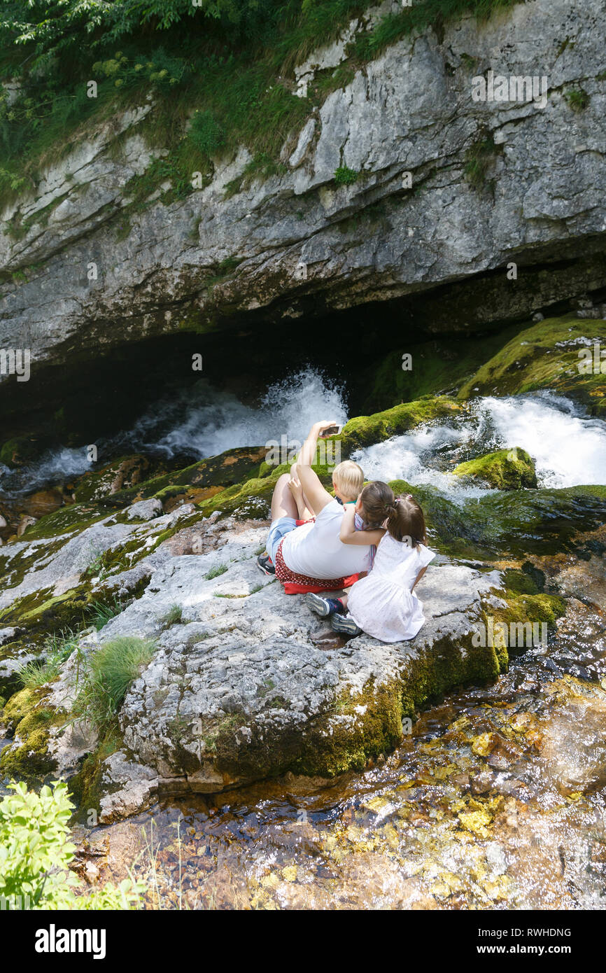 Mutter mit Kindern eine selfie, sitzt auf einem Felsen am Berg Stream auf einer Reise mit der Familie. Lebensstil im Freien, natürlichen Parenting, Kindheit Erfahrung Stockfoto