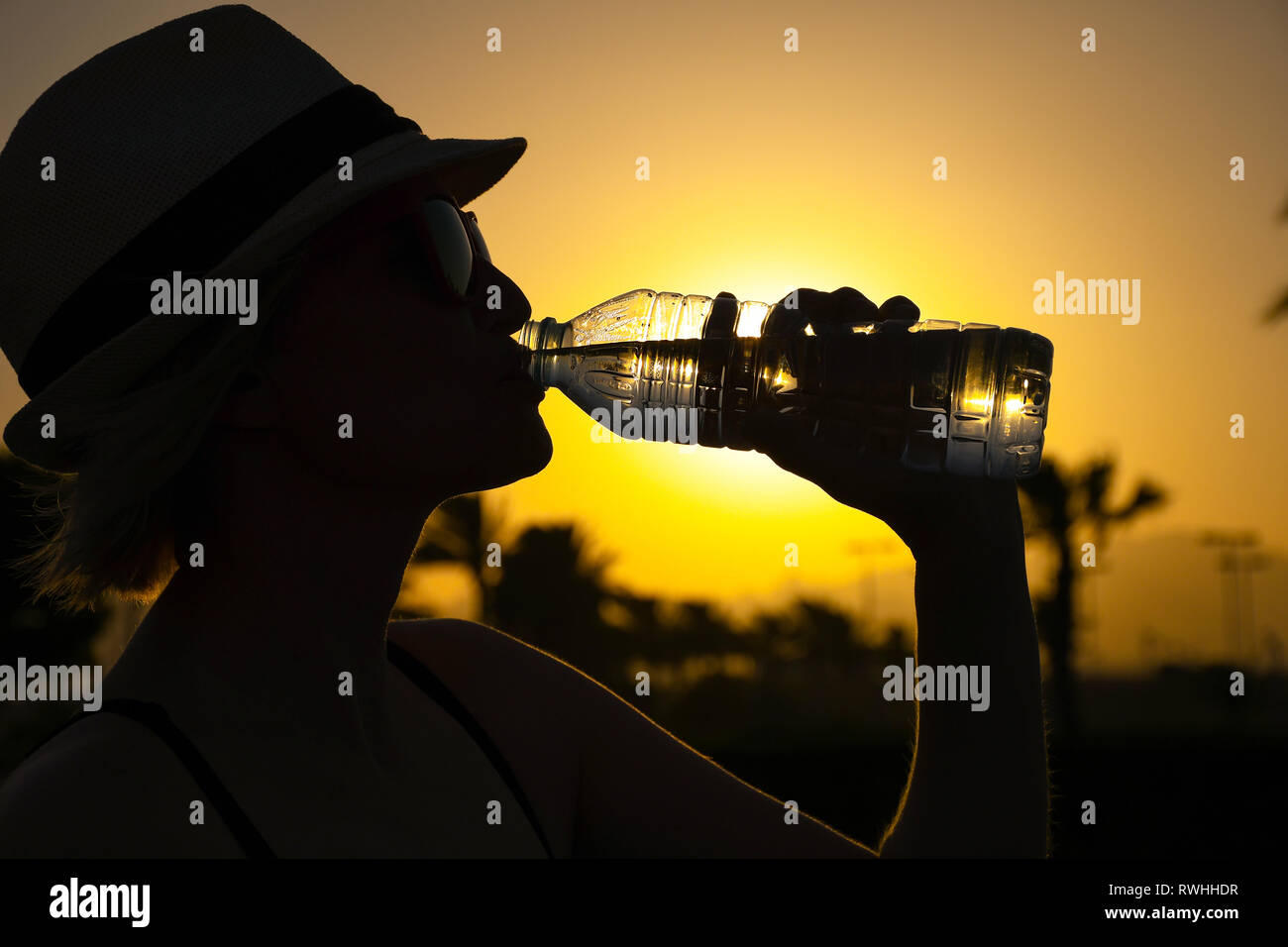 Frau mit weißen Hut und rosa Sonnenbrille mit schönen Reflexion von Palmen und Sonnenuntergang trinken Frisches reines Wasser außerhalb an einem sonnigen Tag aus einem Glas Stockfoto