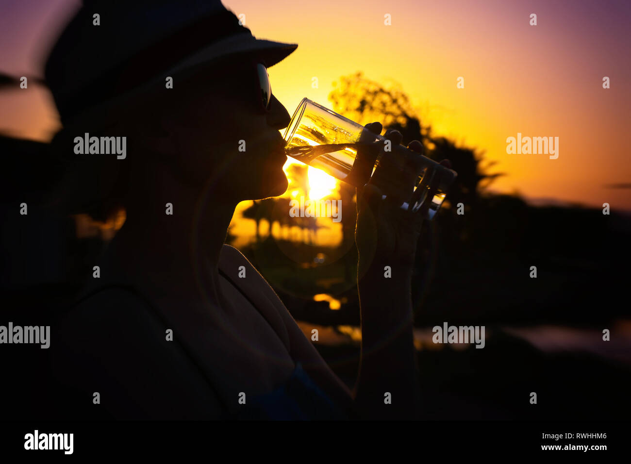 Frau mit weißen Hut und rosa Sonnenbrille mit schönen Reflexion von Palmen und Sonnenuntergang trinken Frisches reines Wasser außerhalb an einem sonnigen Tag aus einem Glas Stockfoto