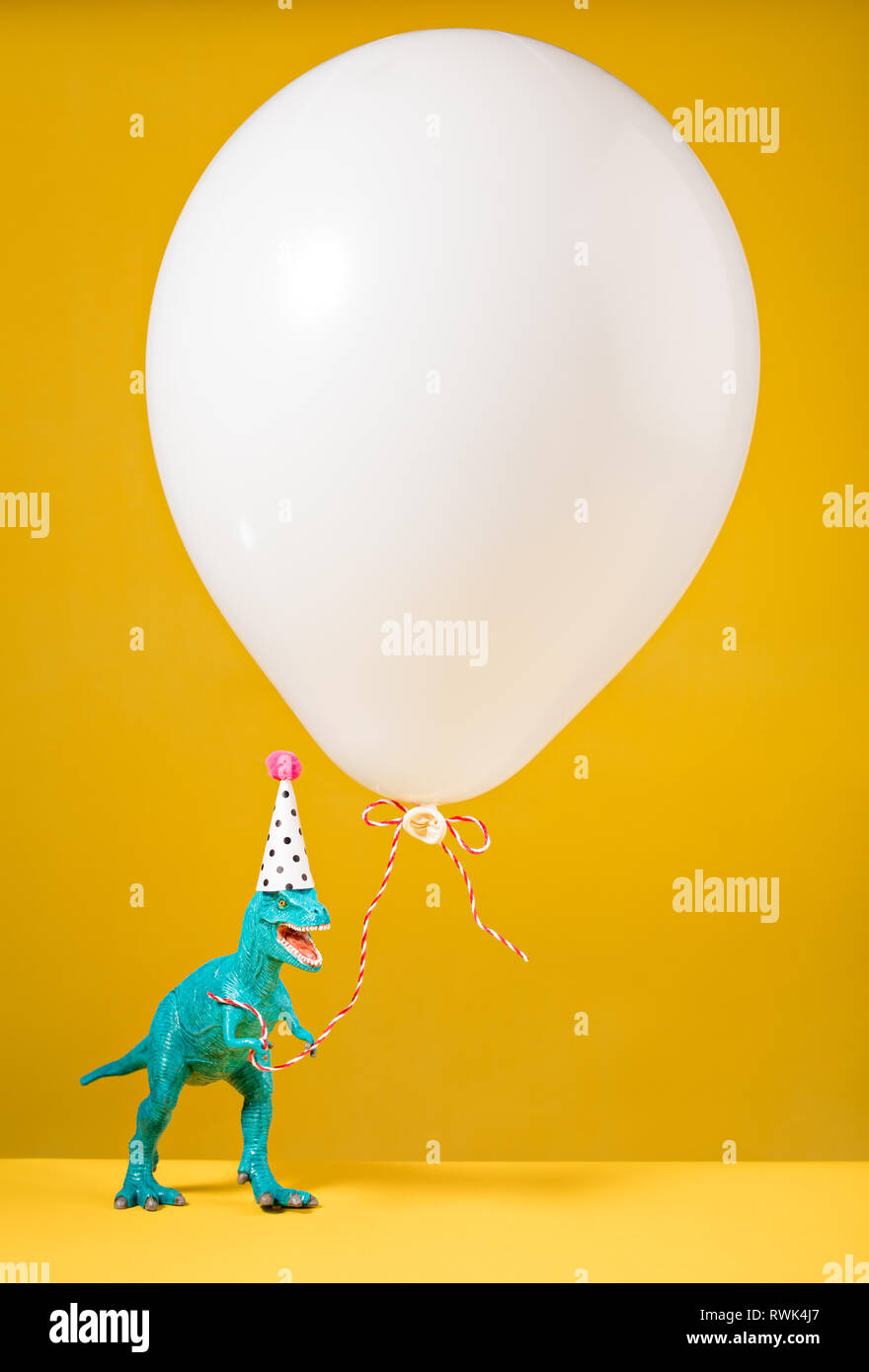 Teal Dinosaurier Spielzeug mit geburtstagsmütze Holding ein weisser Ballon auf gelbem Hintergrund. Stockfoto