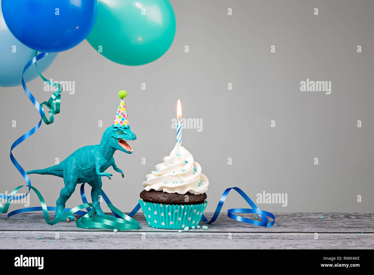Blauer Dinosaurier Spielzeug mit Geburtstag Kuchen und Luftballons auf grauem Hintergrund. Stockfoto