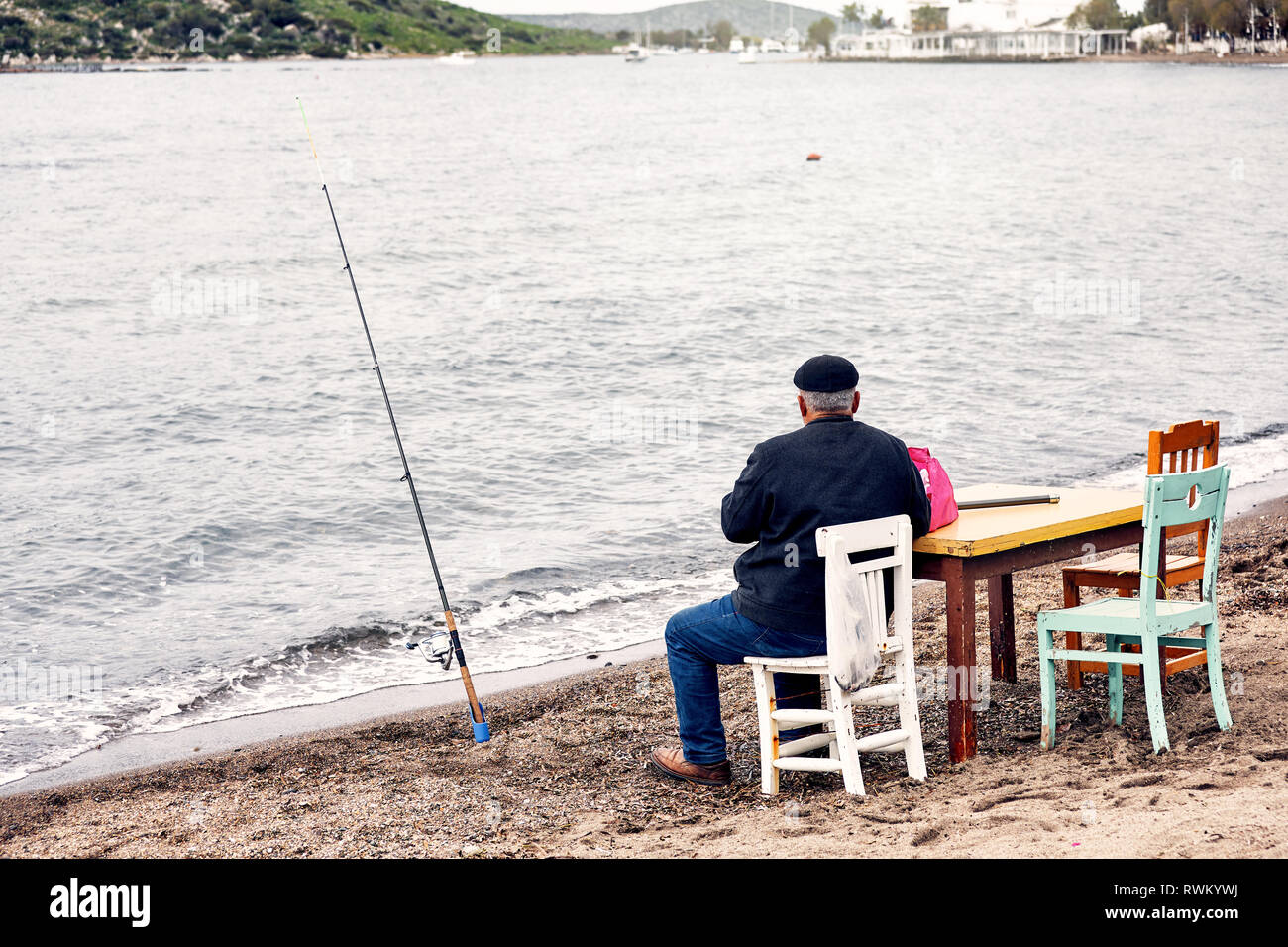 Alte einheimische türkische angler Angler sitzen auf einem hölzernen Stuhl und Angeln in der Nähe der Küste an einem trüben Wintertag. Stockfoto