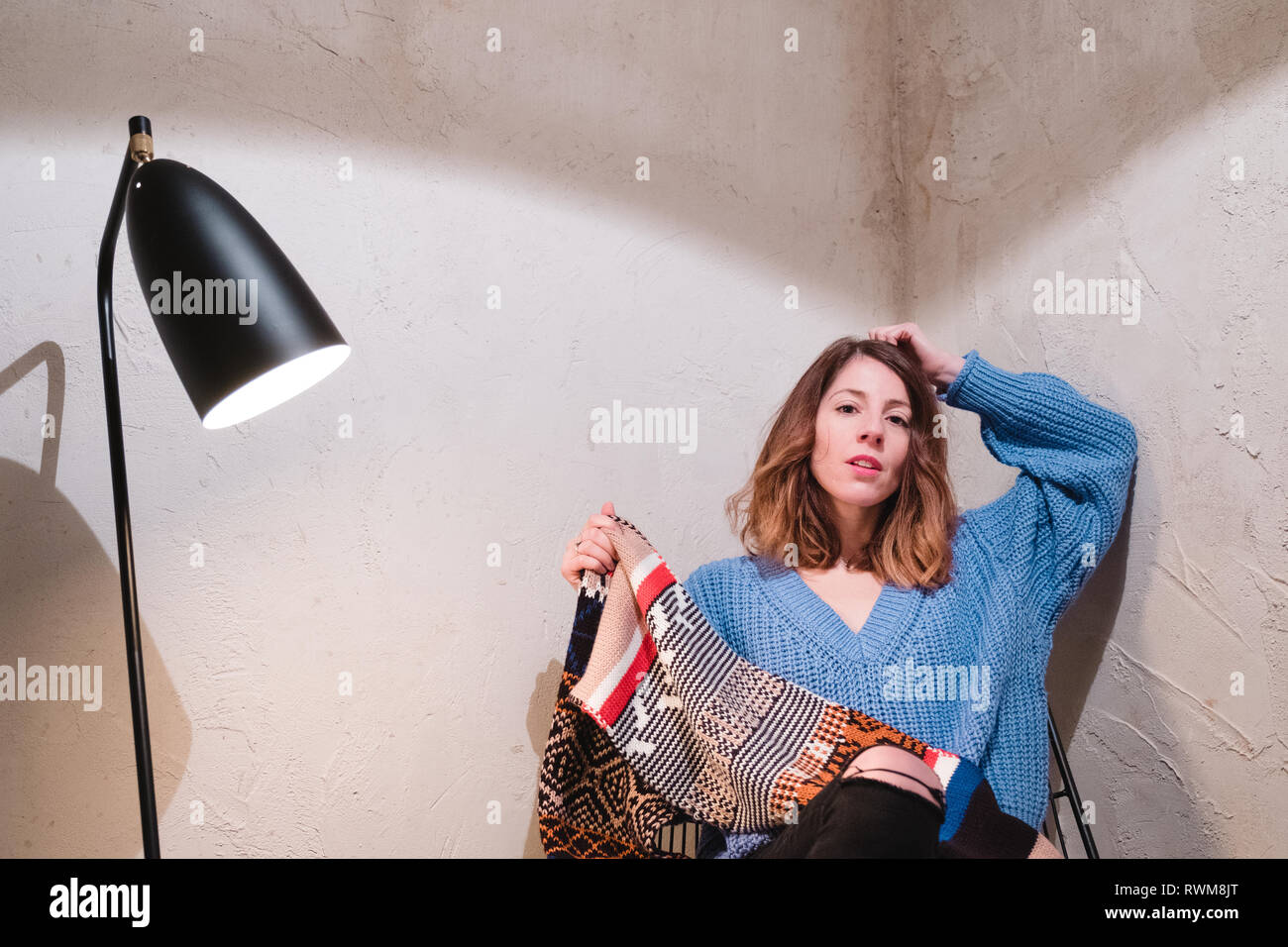 Junge Dame in Form gestrickter Pullover mit Schal sitzen auf Stuhl in der Nähe von Mauer und Lampe im Zimmer Stockfoto
