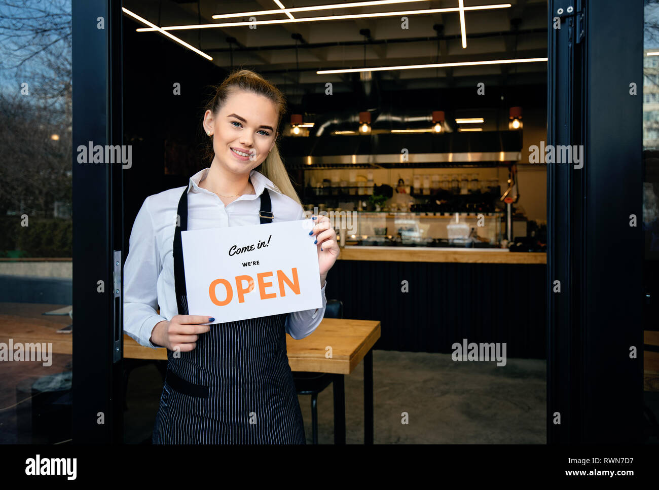 Kellnerin tragen Schürze stehen im Restaurant Eingang. Frau teilnehmen, neue Kunden in ihrem Café. Glückliche Frau, offene Zeichen in Ihrem kleinen Busi Stockfoto