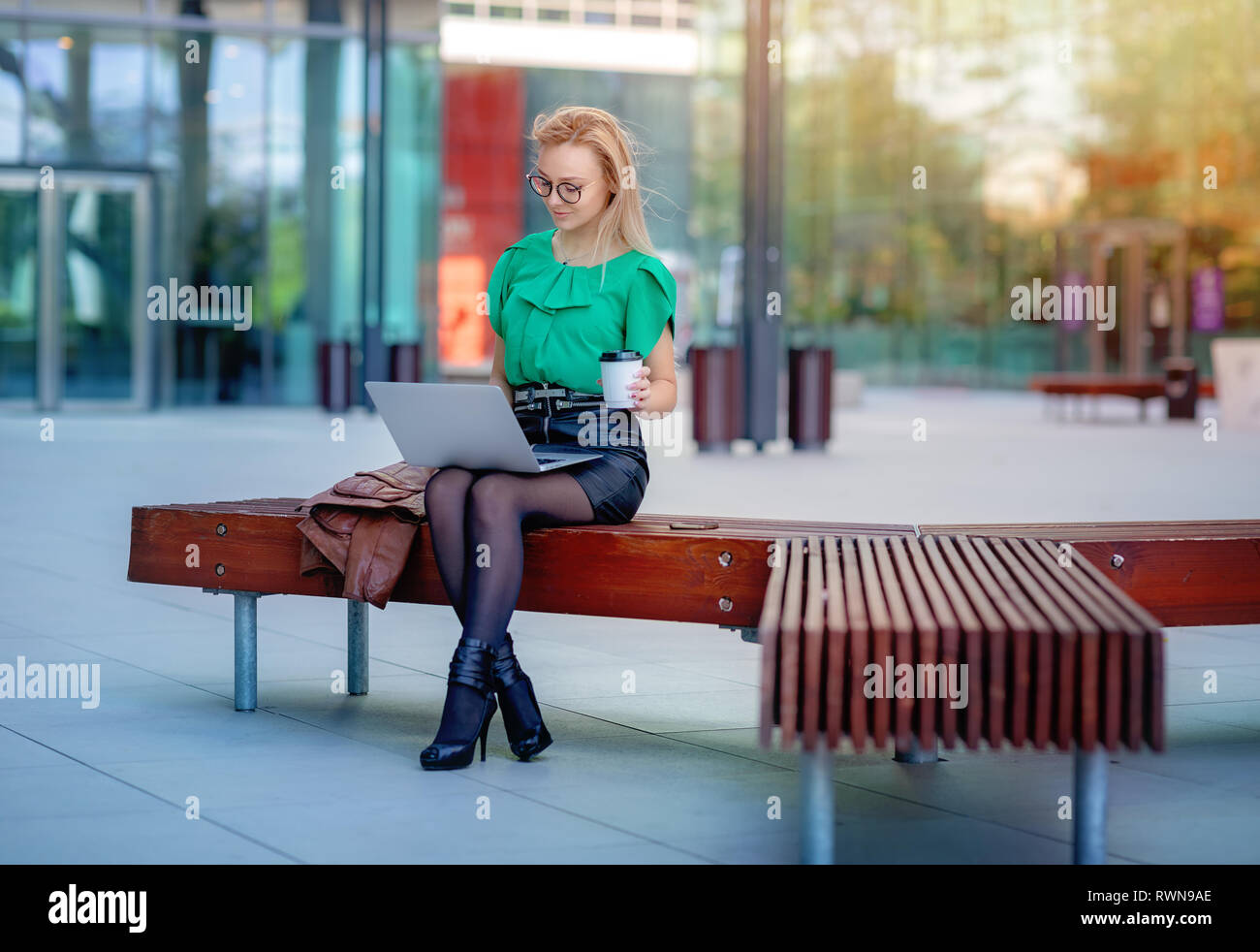 Junge Geschäftsfrau mit einem Laptop und trinken Kaffee auf einer Bank außerhalb des Bürogebäudes. Moderne Bürogebäude Stockfoto