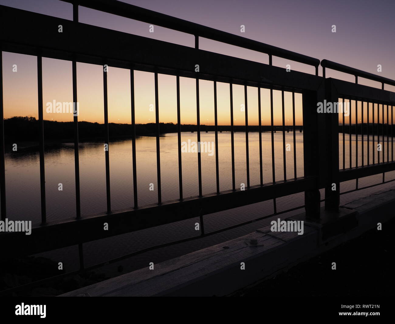 Ein Sonnenuntergang Silhouette einer Brücke über einen Fluss. Schönen farbigen Sonnenuntergang mit markante Silhouette der vertikalen Linien aus Handläufe zu den Fluss suchen Stockfoto