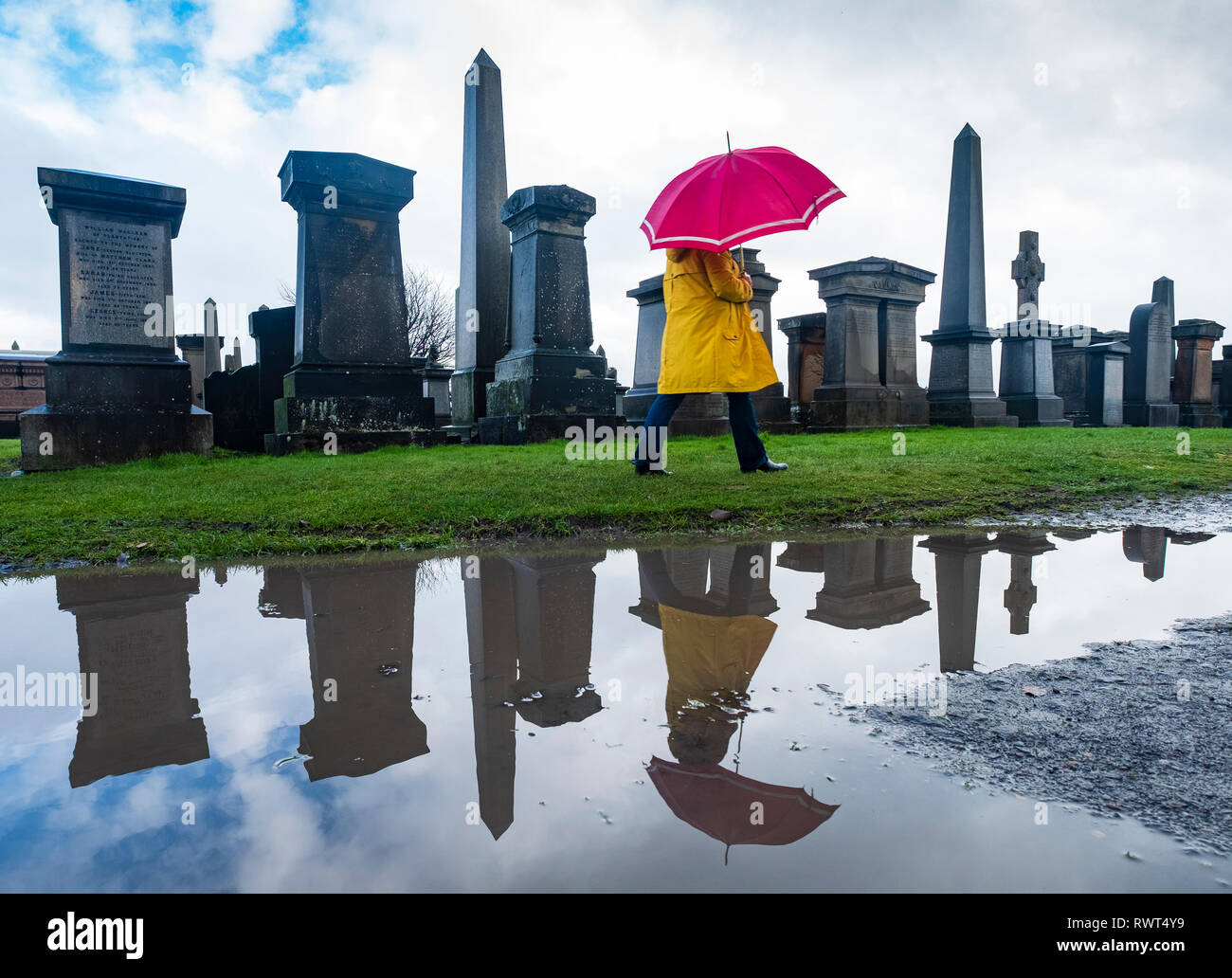 Frau in Gelb und Rot Regenschirm in Pfütze wandern in Nekropole Friedhof in Glasgow, Schottland, Großbritannien wider Stockfoto