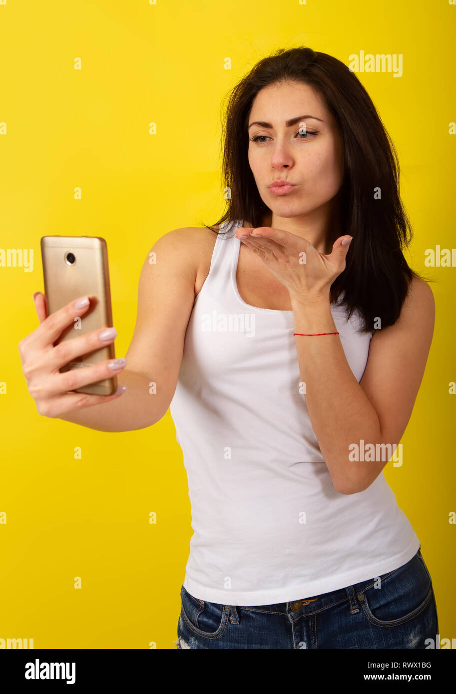 Charmante junge Dunkelhaarige in modischen legere Kleidung macht einen selfie am Telefon und Grimassen auf einem hellen gelben Hintergrund Stockfoto