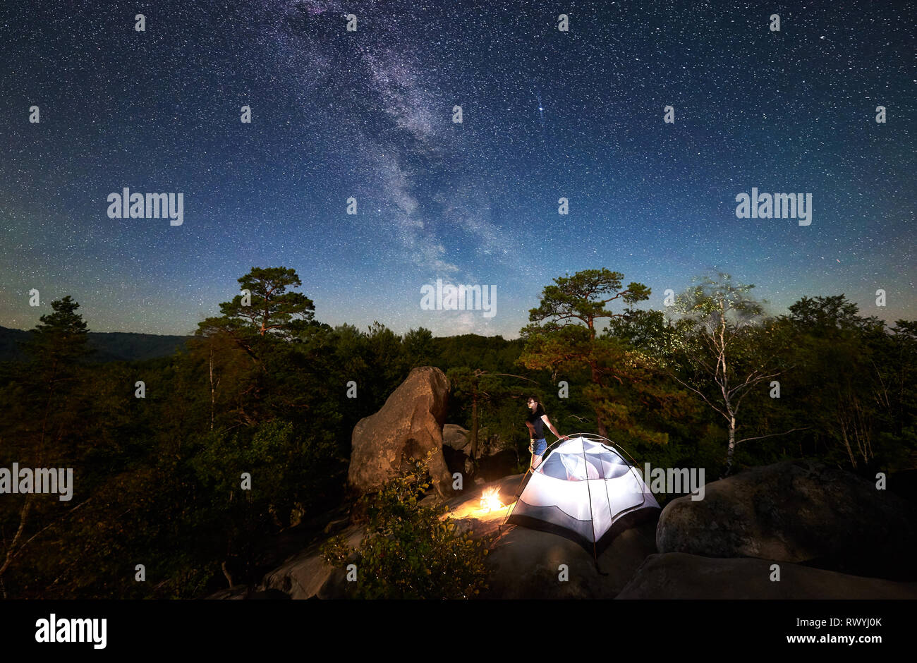 Frau entspannend auf Rocky Mountain Top neben Camp, Bonfire und touristische Hütte im Sommer Nacht, mit Sicht auf den Himmel voller Sterne und Milchstraße. Auf Hintergrund Sternenhimmel, große Steine und Bäume Stockfoto