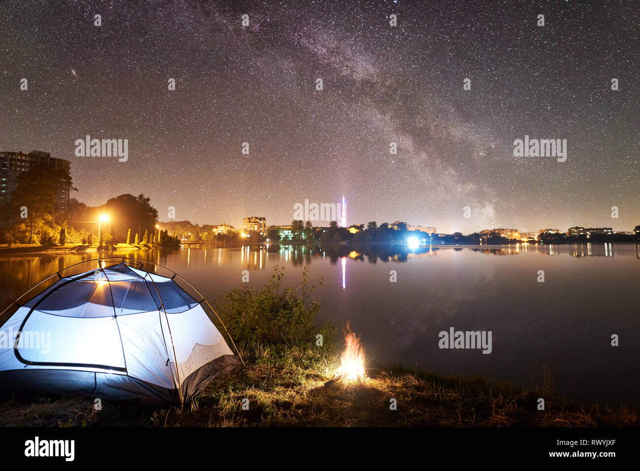 Nacht Camping. Beleuchtete touristische Zelt und Lagerfeuer auf dem Lake Shore unter schönen Abend Himmel voller Sterne und die Milchstraße, ruhige Wasseroberfläche, leuchtende Stadt auf Hintergrund. Outdoor Lifestyle Konzept Stockfoto