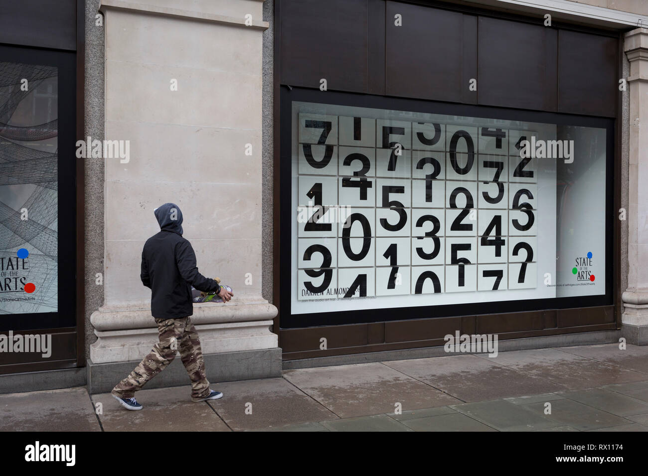 Ein Skateboarder Spaziergänge Vergangenheit ein Fenster angezeigt, in dem Sie mit Zahlen - Teil eines Designs Thema 'State der Künste" genannt, im Kaufhaus Selfridges in der Oxford Street, am 4. März 2019 in London, England. Darren Almonds Stück "Zufällige Begegnung 004", besteht aus einem Gitter aus rechteckigen Gremien gebildet, mit fragmentierten Zahlen, die scheinbar auf der Oberfläche zu blättern. Zustand der Kunst ist eine Galerie mit Werken von neun crtically-anerkannten Künstlern in Selfridges, um die Macht der öffentlichen Kunst feiern. Stockfoto