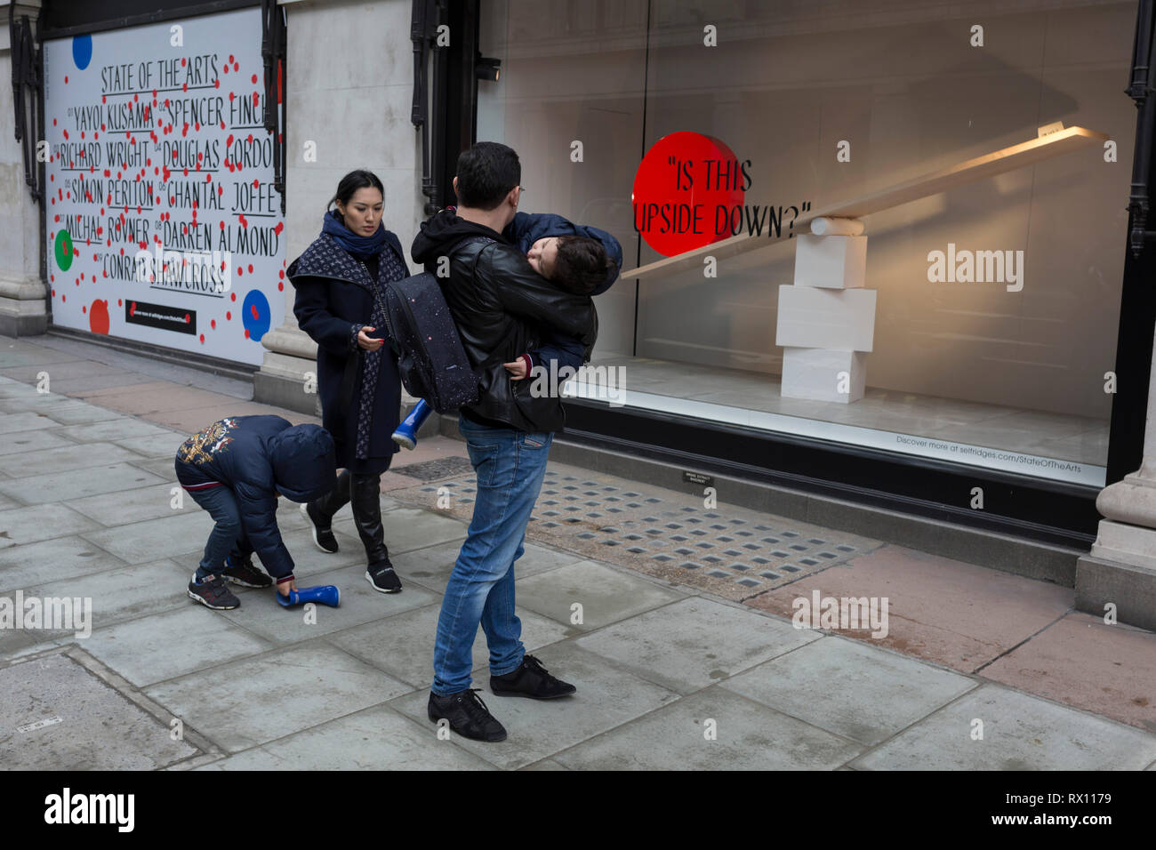 Eine asiatische Familie Kampf mit einem blauen Boot außerhalb eines Fensters angezeigt, die Teil einer Design 'State der Künste" genannt, im Kaufhaus Selfridges in der Oxford Street, am 4. März 2019 in London, England. Zustand der Kunst ist eine Galerie mit Werken von neun crtically-anerkannten Künstlern in Selfridges, um die Macht der öffentlichen Kunst feiern Jeder der Künstler sind in der Erstellung einer Site-spezifischen Kunstwerk an einem der neuen Elizabeth Linie Stationen als Teil der Traverse Art Programm beteiligt. Stockfoto