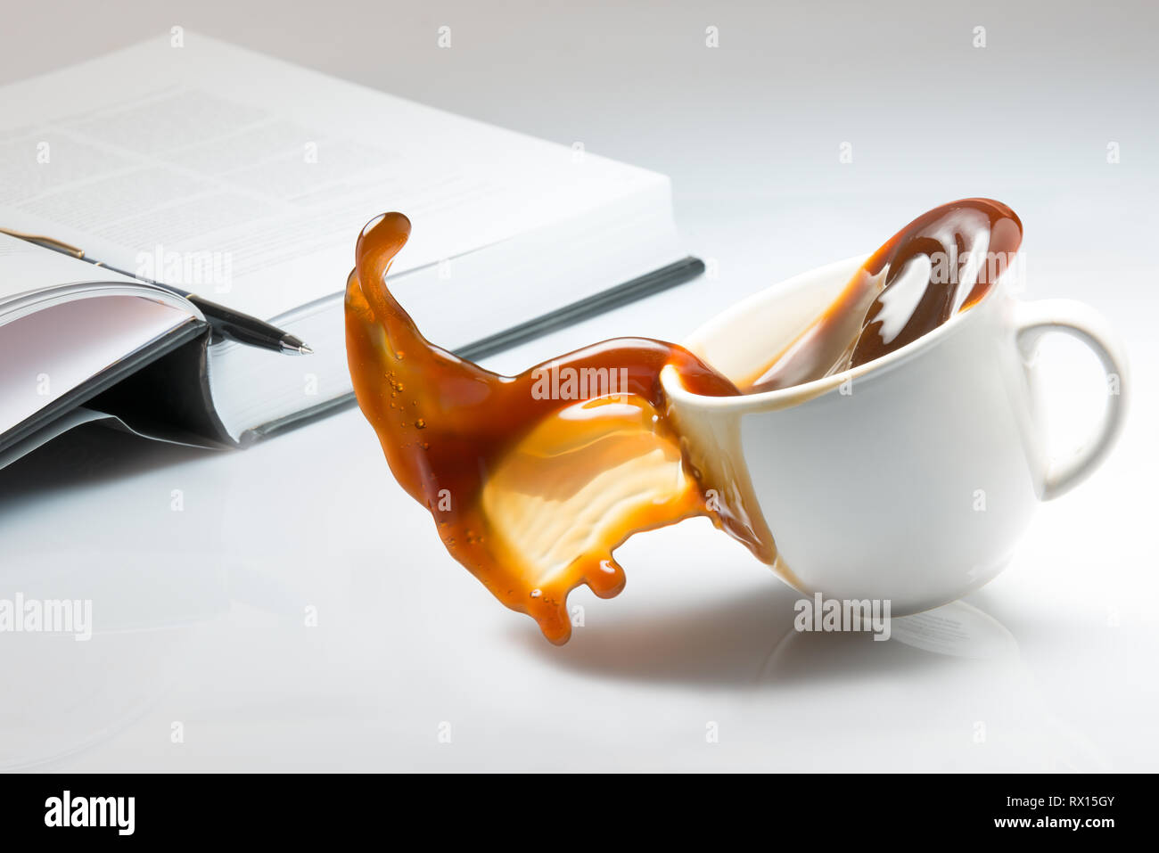 Fallende und Verschütten von einer Tasse Kaffee auf einem Schreibtisch mit einem offenen Buch Stockfoto
