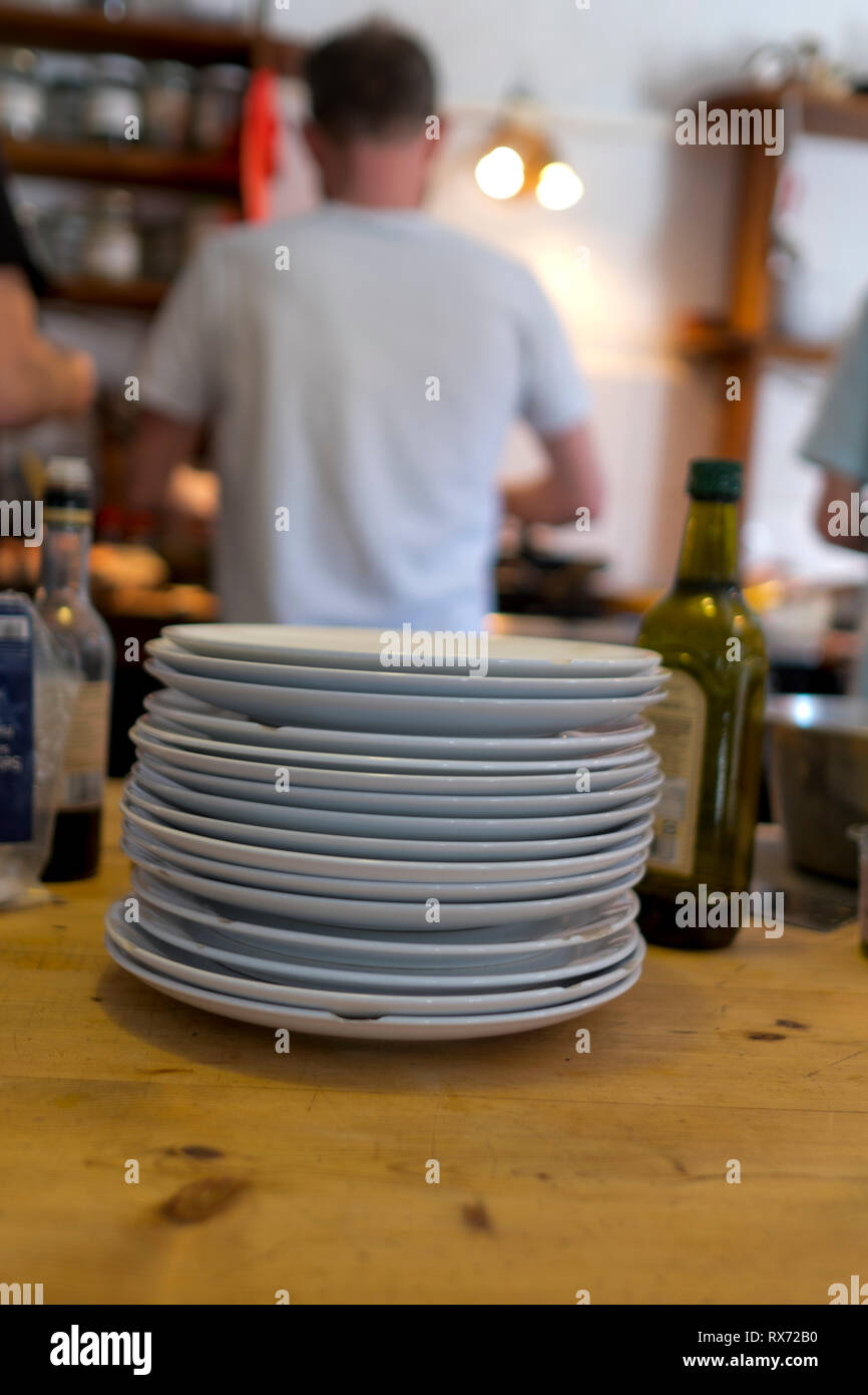 Ein Stapel von abgebrochenem weißen Platten sitzen auf einem hölzernen Kieferntisch im Vordergrund in der Küche mit drei Männern Kochen im Hintergrund Stockfoto