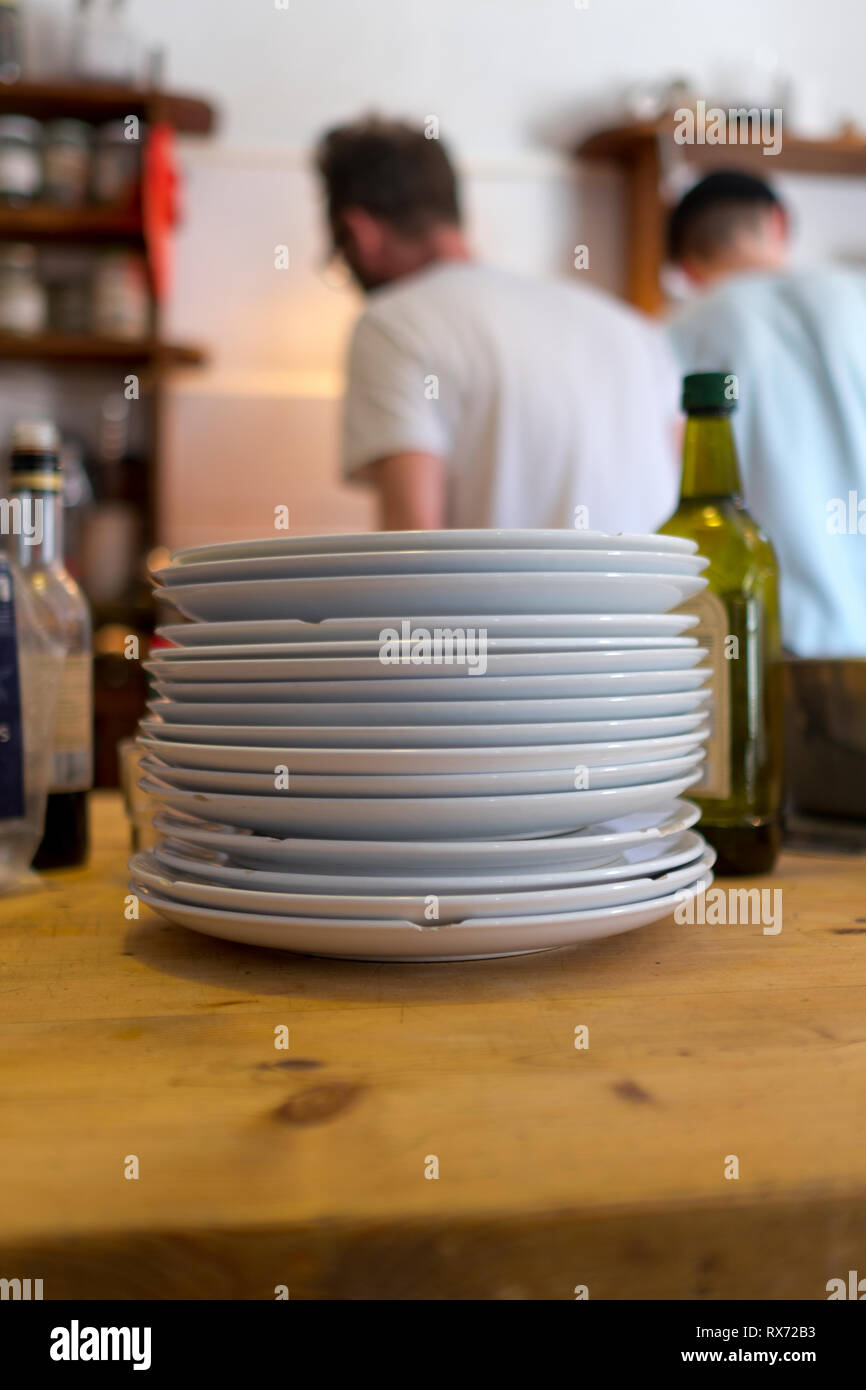 Ein Stapel von abgebrochenem weißen Platten sitzen auf einem hölzernen Kieferntisch im Vordergrund in der Küche mit zwei Männern Kochen im Hintergrund Stockfoto