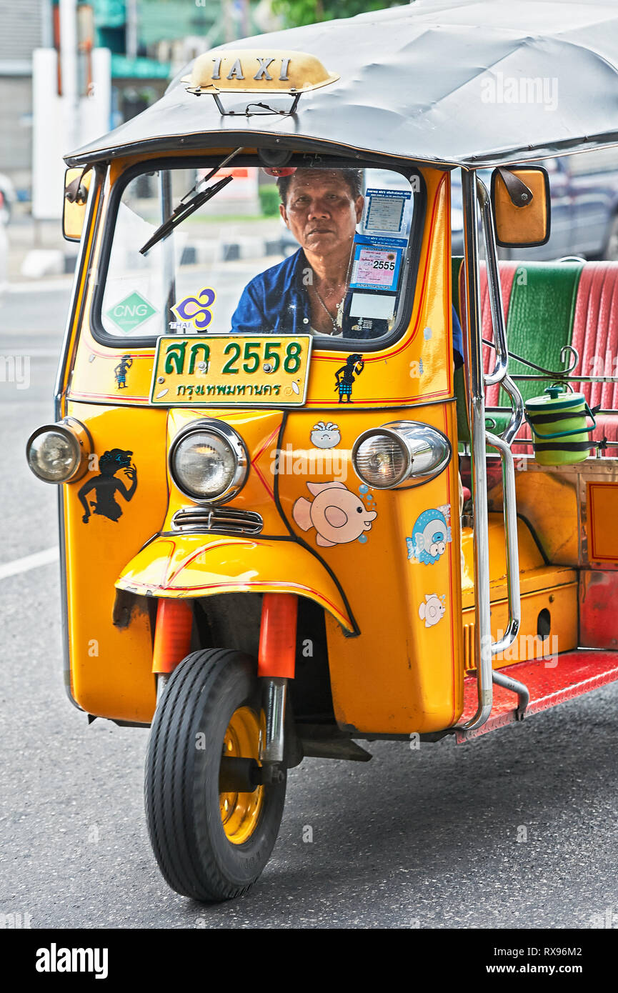 Bangkok, Thailand: Close-up Vorderansicht eines Gelb lackiert Tuk-tuk mit einem mittlerem Alter männliche Fahrer, von dem übrigen Datenverkehr isoliert Stockfoto