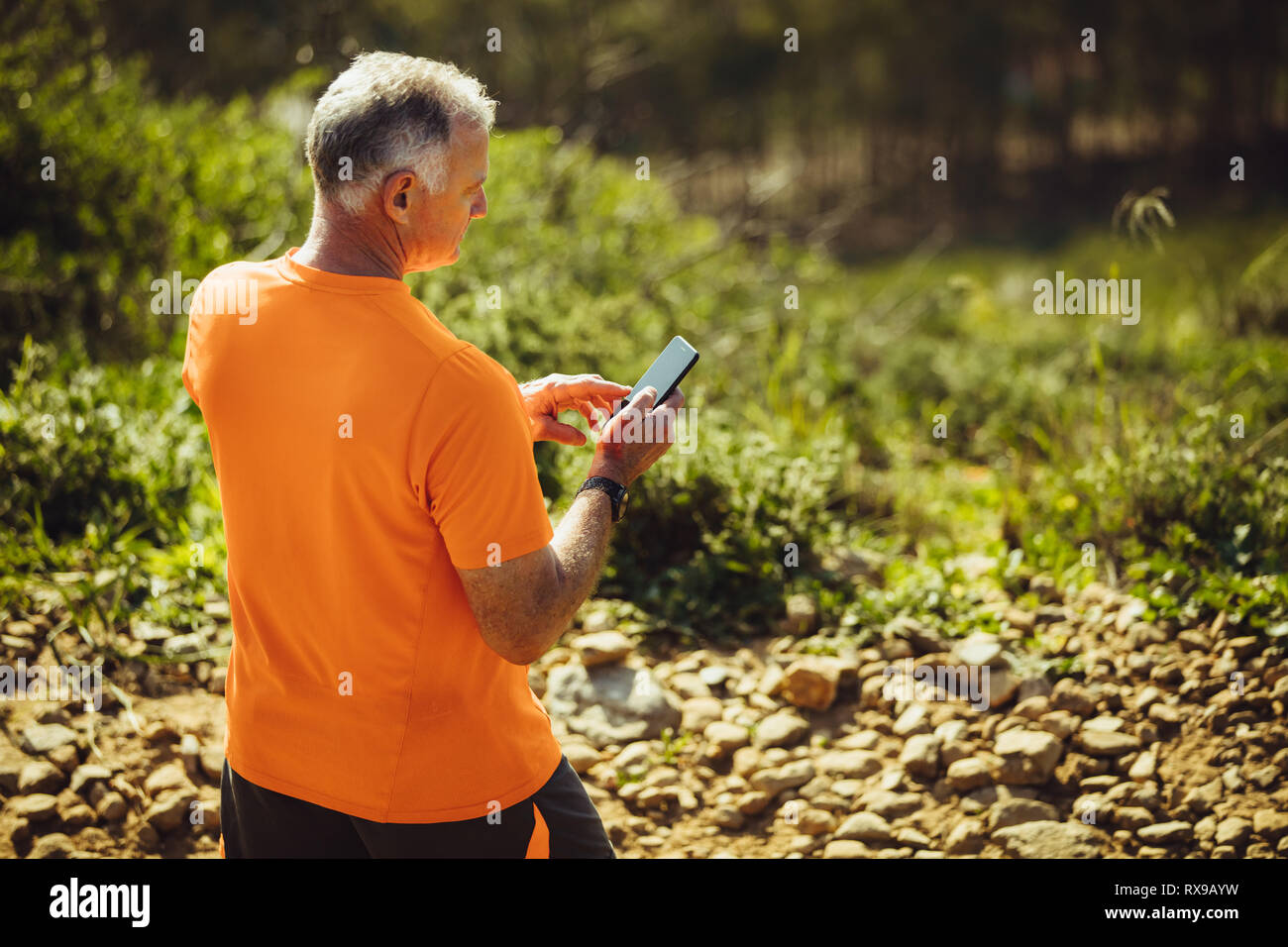 Ansicht von hinten von einem Mann an seinem Telefon beim Gehen auf einem felsigen Weg weg. Älterer Mann in Fitness Tragen Sie wandern auf einem Gelände an einem sonnigen Tag. Stockfoto
