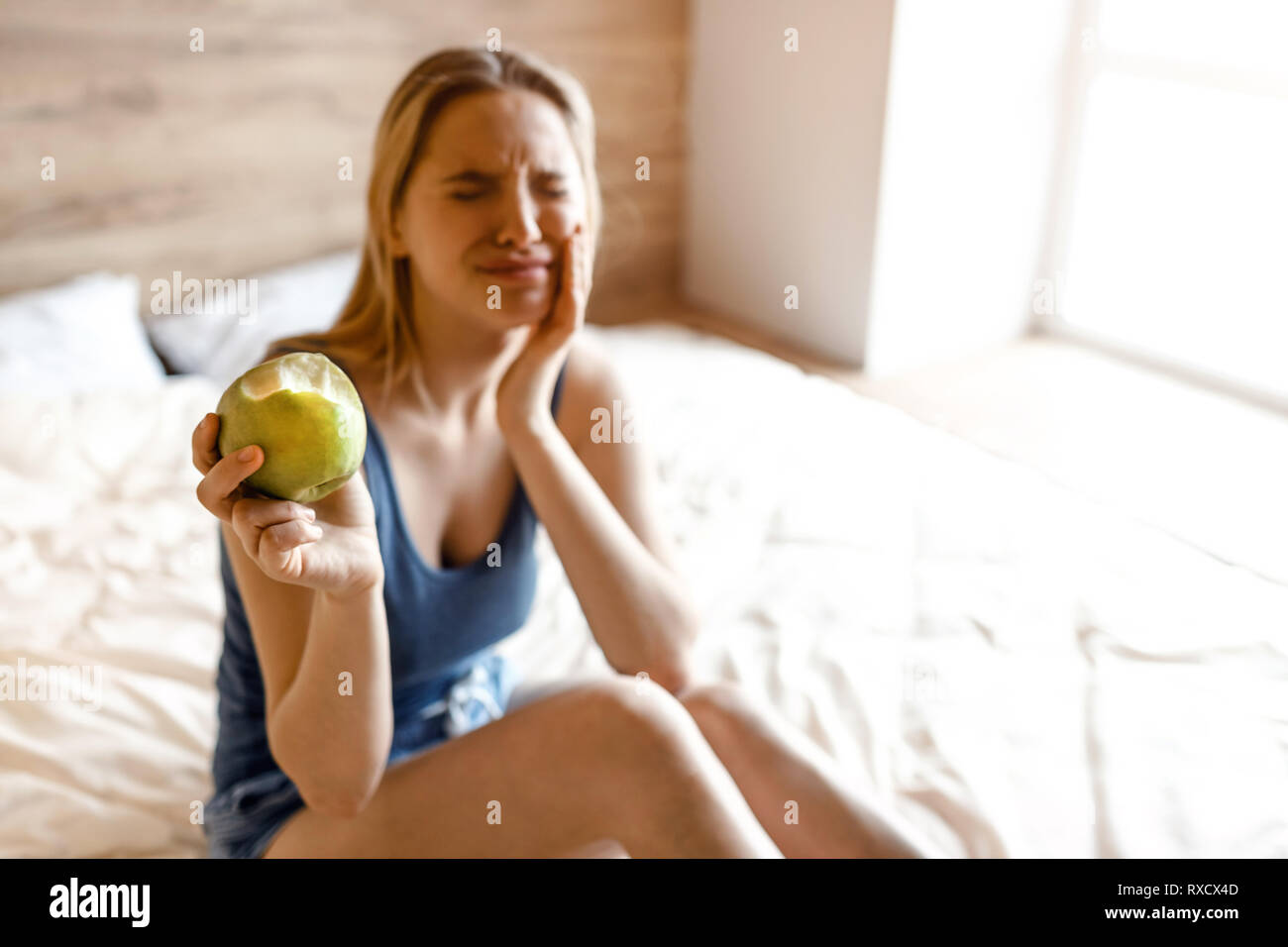 Junge hübsche blonde Frau im Bett am Morgen sitzen. Modell leiden Zahn weh. Sie halten die Hand auf die Wange. Gebissen Apfel in der anderen Hand. Sie zeigen Ich Stockfoto