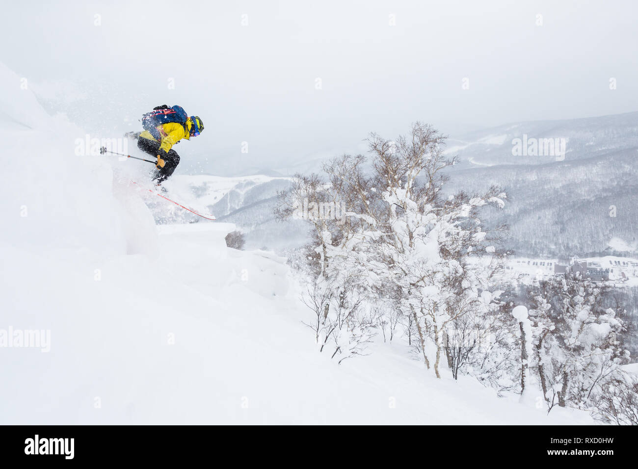Backcountry skier springt von einem Gesims in der Nähe der Gipfel eines kleinen Spitze im backcountry Hokkaido, Japan. Gelbe Jacke, blau Rucksack Tiefschneefahren in Ja Stockfoto