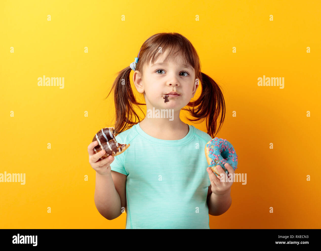 4-Jähriges Mädchen in einem Türkis t-shirt Essen Donuts. die Haare des Mädchens in Schwänze gebunden ist. Orange hinterlegt. Stockfoto