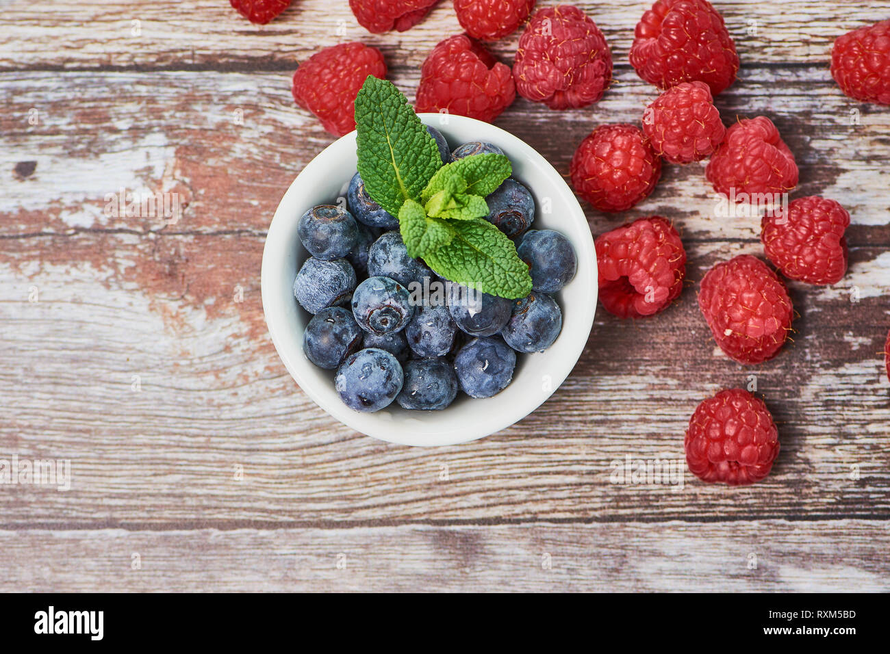 Draufsicht auf Heidelbeeren in einer weissen Schüssel mit etwas Pfefferminze in der Mitte eines Holz- Hintergrund, einige Himbeeren in der Ecke Stockfoto