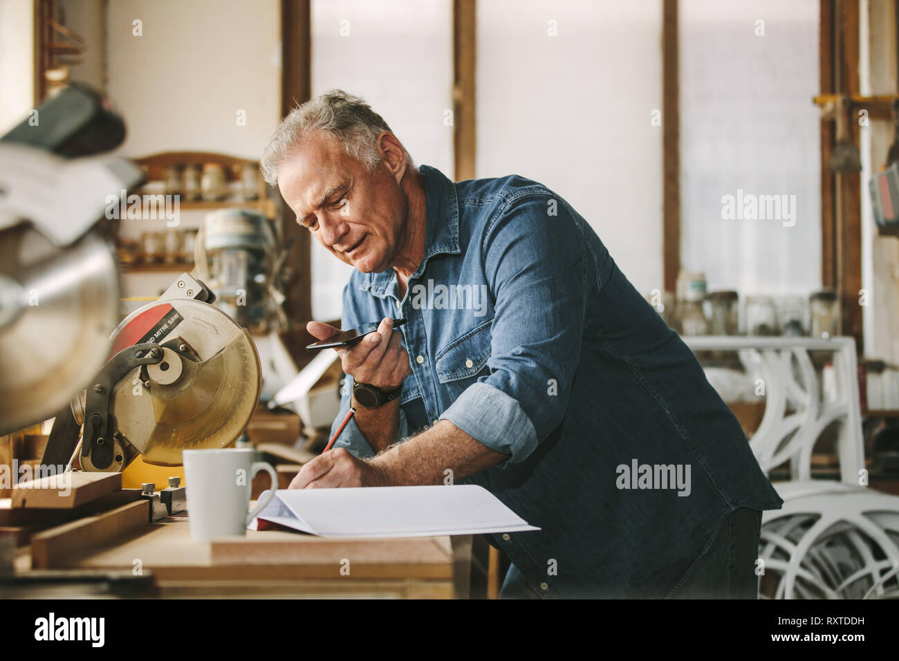 Senior Zimmermann in seiner Werkstatt arbeiten am Schreibtisch mit Handy stehen und Schreiben in einem Buch. Reife Männer arbeiten in der Schreinerei. Stockfoto