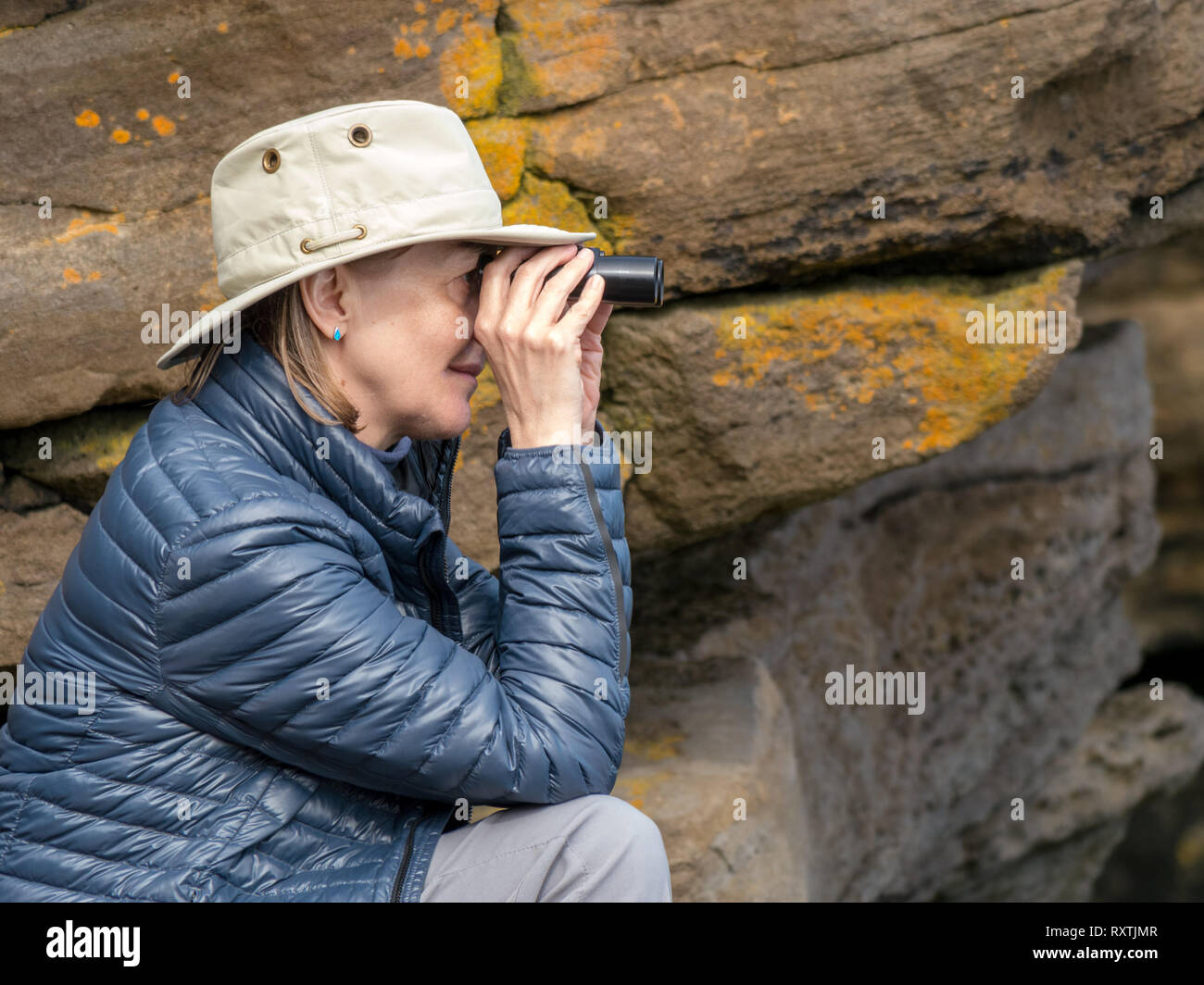 Erwachsene Frau mit Sonnenhut und gepolsterter Pufferjacke, die durch ein Fernglas schaut und wilde Tiere mit felsigen Klippen dahinter beobachtet, Isle of Skye, Schottland, Großbritannien Stockfoto