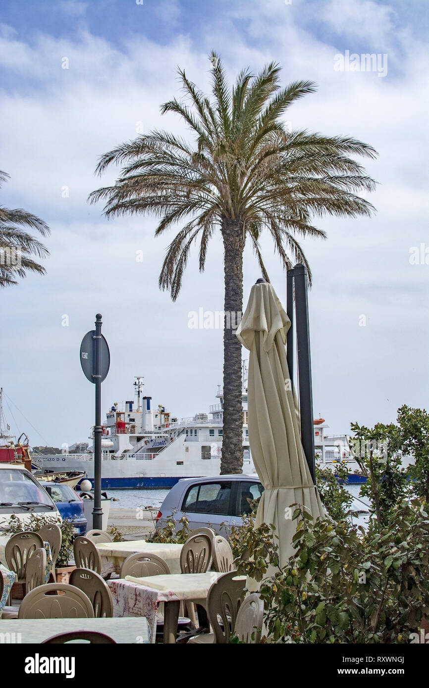 ISOLA LA MADDALENA, Sardinien, Italien - 7. MÄRZ 2019: Fähre in den Hafen über Cafe Tische im Freien mit Palme an einem bewölkten Tag am 7. März 2019 in La M Stockfoto