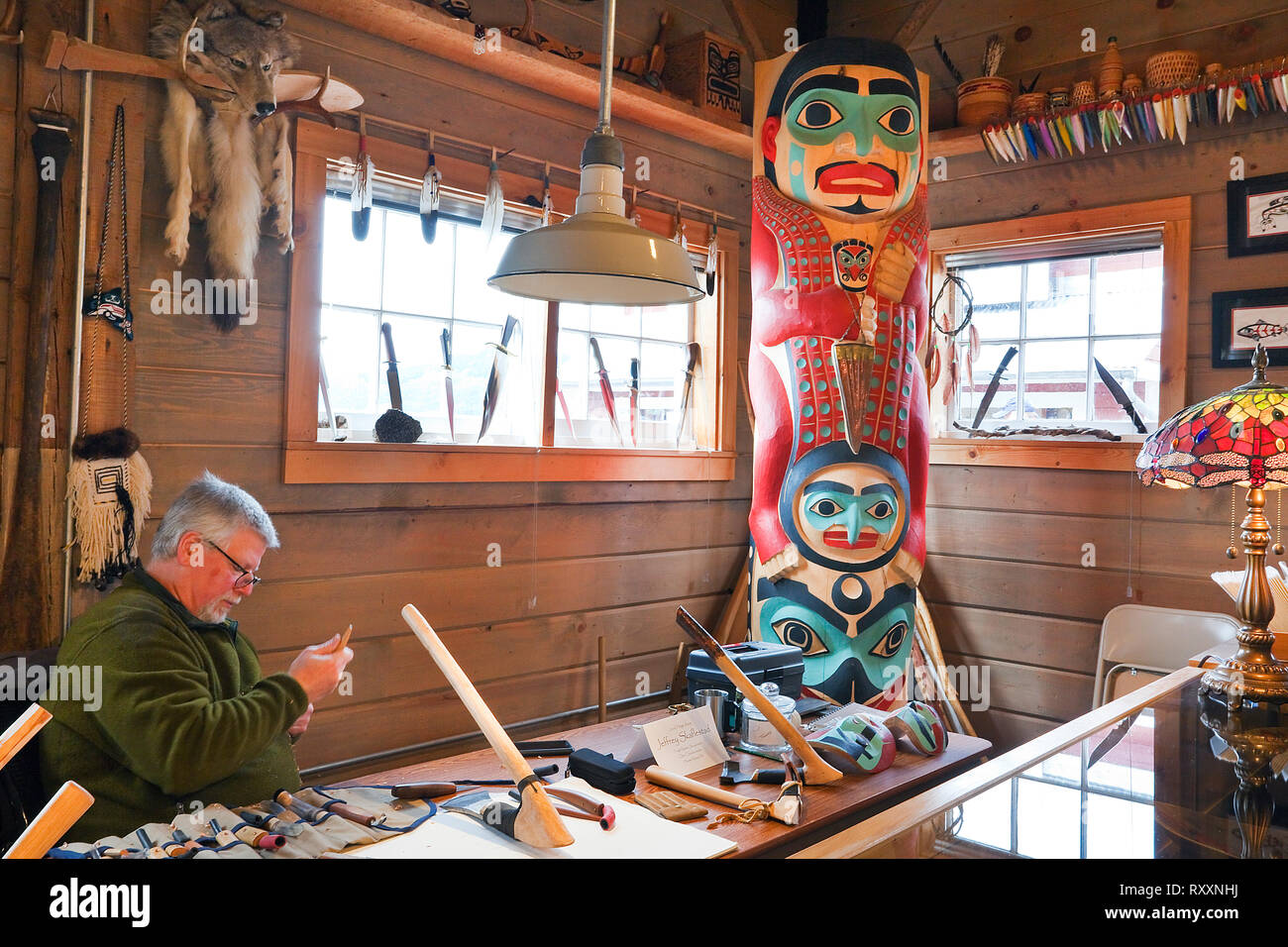 Norwegisch-Tinglit-Schnitzer Jeff Skaflestad an einem Arbeitstisch im Icy Strait Point Cannery Museum. Das Totem in der Ecke trägt den Titel 'Hawkt' Tooth Warrior'. ICY Strait Point, Alaska, USA Stockfoto