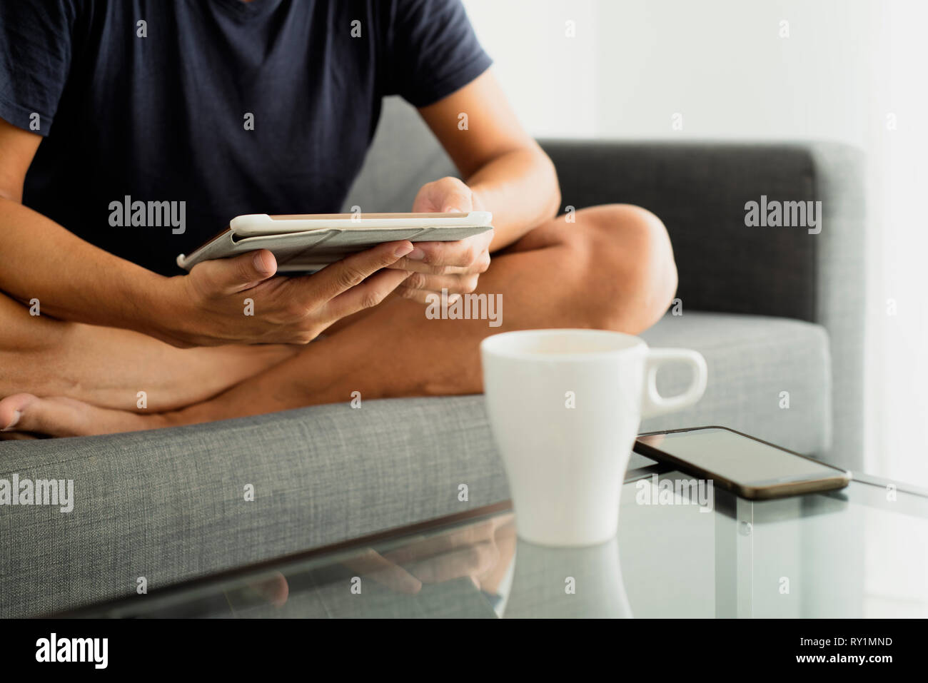 Nahaufnahme eines jungen kaukasischen Mann in kurzen Hosen sitzen im Schneidersitz, in einem Sofa, und wird über eine Tafel und eine Tasse Kaffee und ein Smartphone auf einem Glas c Stockfoto