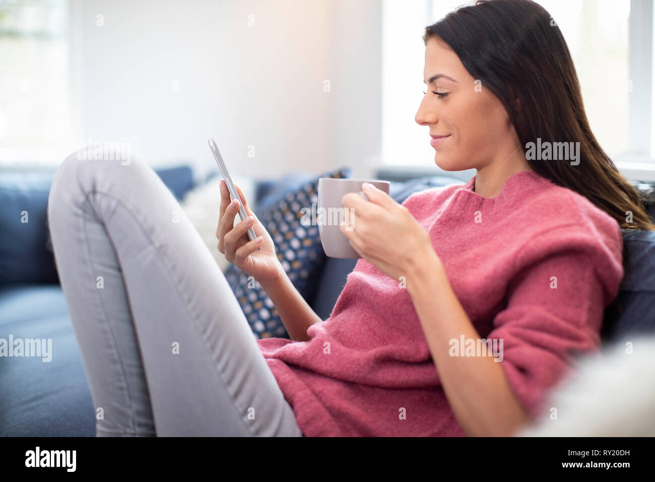 Junge Frau auf Sofa mit heißen Getränken Kontrolle Mobiltelefon Stockfoto