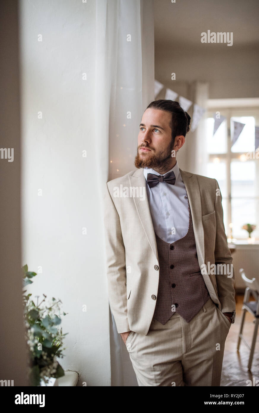 Ein schöner Hipster jungen Mann oder ein Bräutigam mit Anzug stehen durch die Fenster auf der einen Partei, die Hände in den Taschen. Stockfoto
