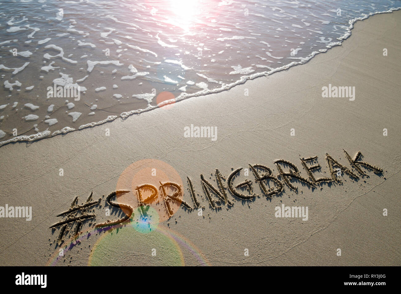 Moderne Spring Break Nachricht handschriftlich mit einem social media Hashtag auf der glatten Sand eines leeren Strand mit einem entgegenkommenden Wave Stockfoto