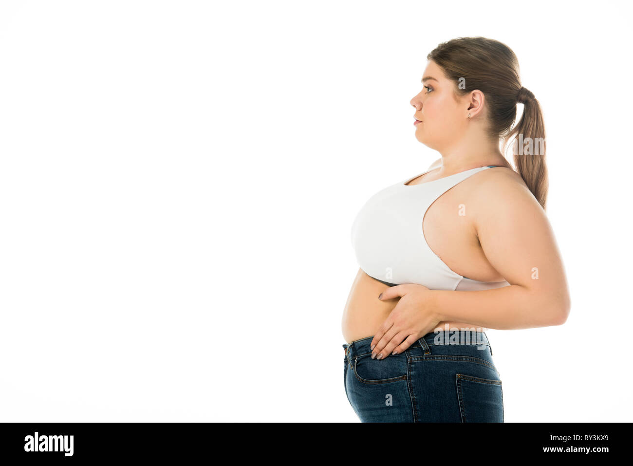 Seitenansicht der übergewichtigen Frau in Denim mit der Hand auf dem Bauch auf Weiß isoliert Posing Stockfoto