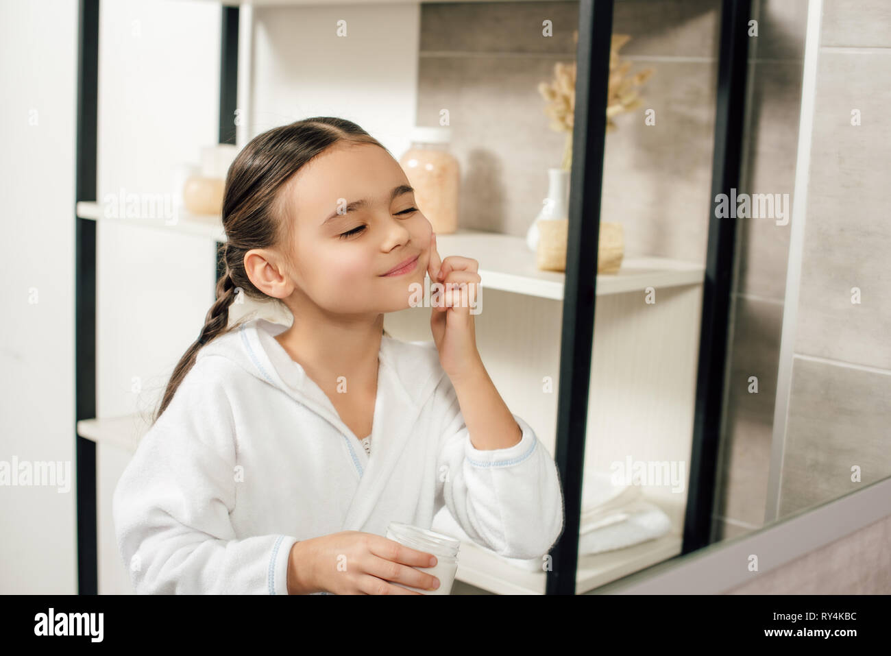 Selektiver Fokus Kind im weißen Bademantel auf der Suche zu spiegeln und Anwendung von kosmetischen Creme im Badezimmer Stockfoto