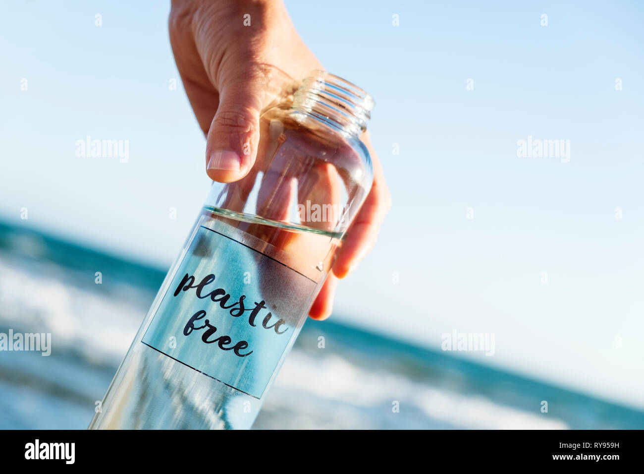 Nahaufnahme von einem kaukasischen Mann hält ein Glas wiederverwendbare Trinkflasche mit dem Text aus Kunststoff in Es geschrieben, am Strand, mit dem Meer in der backgro Stockfoto