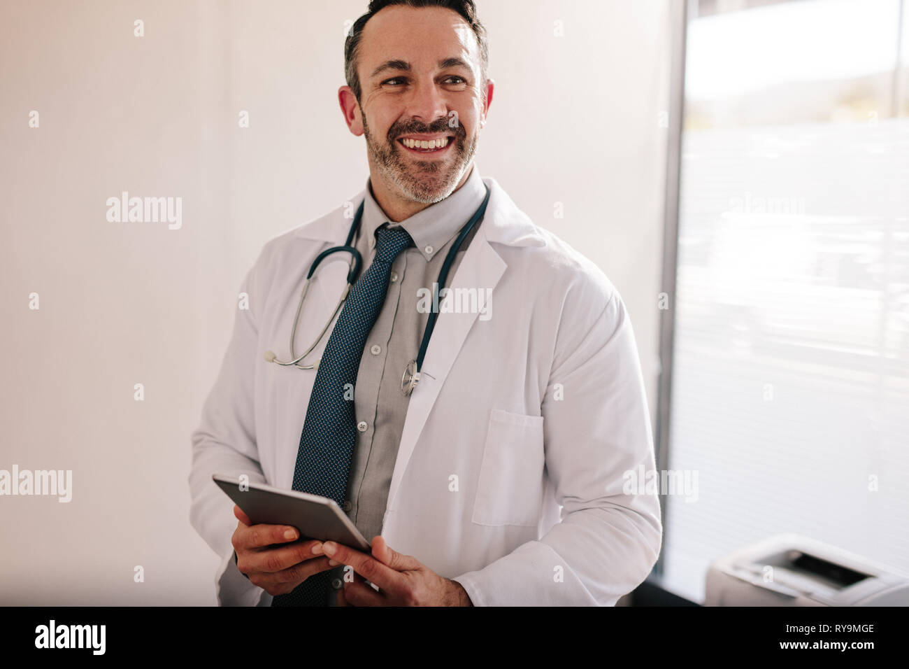 Lächelnd reife männlicher Arzt mit einem digitalen Tablet in seinem Büro. Erfolgreiche medizinische Fachmann an seiner Klinik. Stockfoto