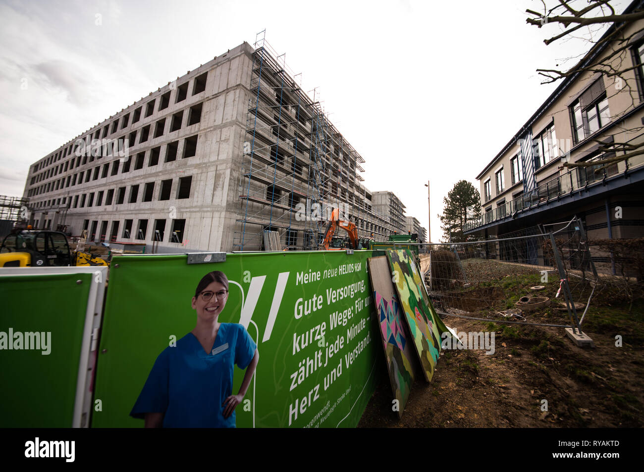 Wiesbaden, Deutschland. 12 Mär, 2019. Poster auf dem Bauzaun über das neue Gebäude des "Dr.-Horst-Schmidt-Klinik' (l Informieren). Der Betreiber, Helios Verwaltung Hessen GmbH, beabsichtigt, das Gebäude bis 2021 abgeschlossen und wird 268 Millionen Euro investieren. Die alte Klinik Gebäude (r) ist von 2023 abgerissen werden. Credit: Andreas Arnold/dpa/Alamy leben Nachrichten Stockfoto