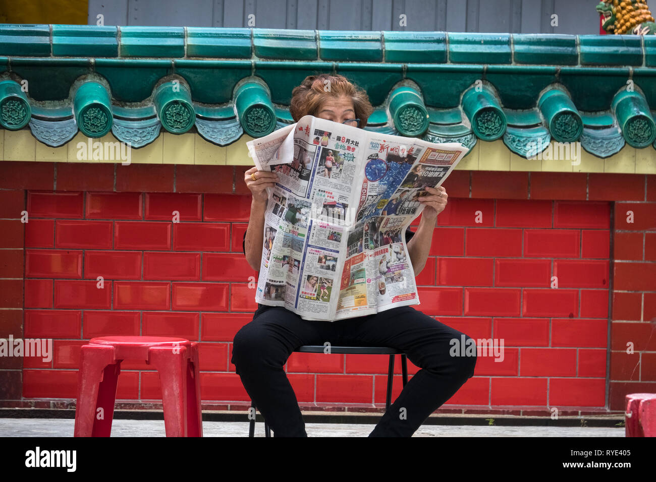 Alte Singapur Frau liest Zeitung auf Stuhl bei chinesischen Tempel - Singapur Stockfoto