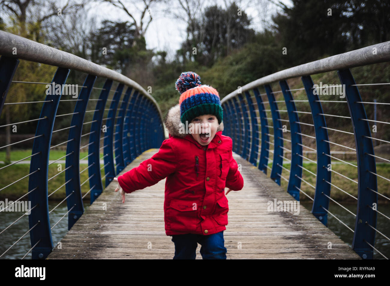 Kleiner Junge auf die Kamera läuft auf Holzbrücke schreien und Spaß haben, trägt einen roten Mantel und Bobble hat Stockfoto