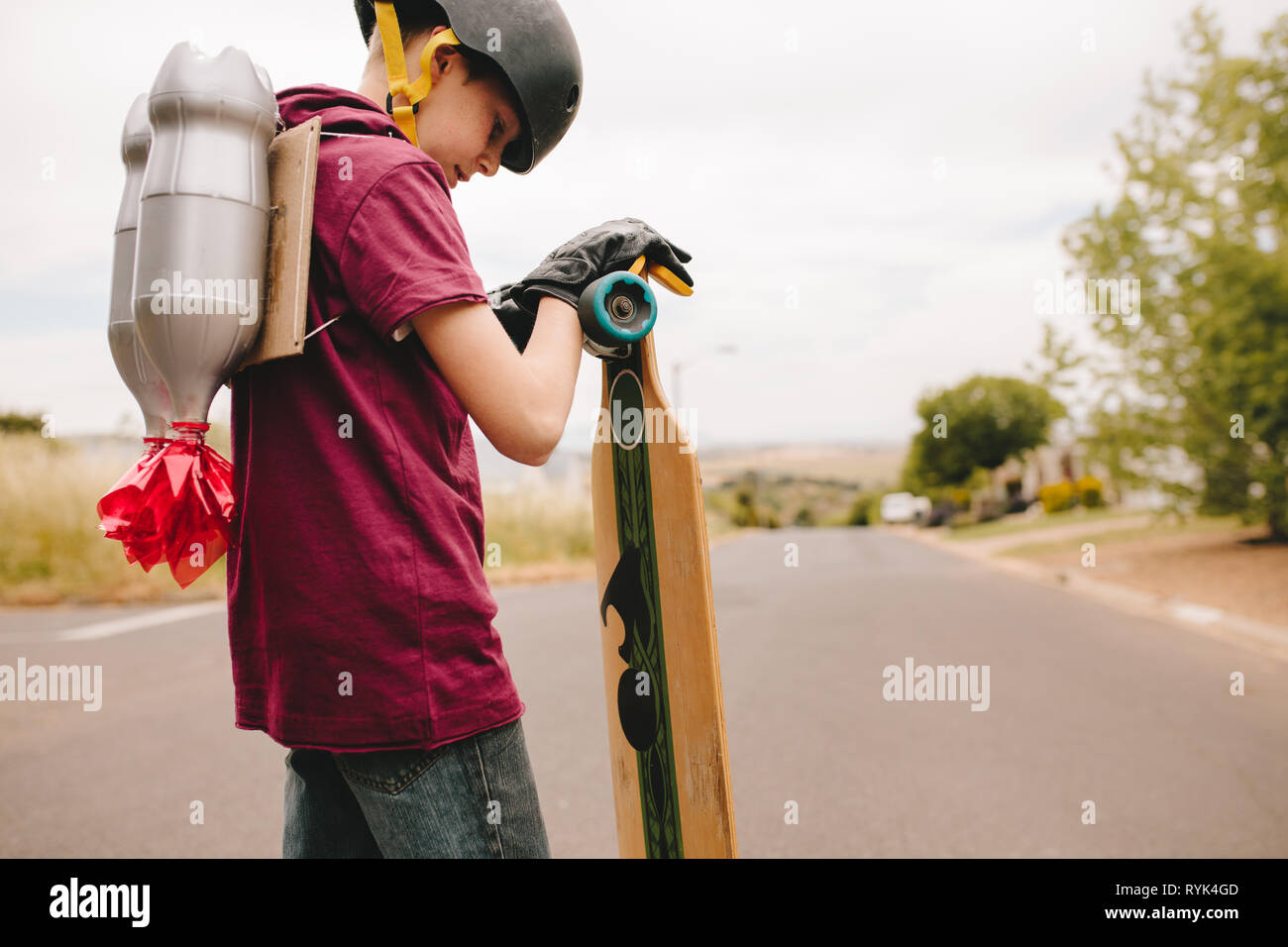 Junge mit Helm mit einem Spielzeug Jetpack auf dem Rücken stehen im Freien mit seinem Skateboard. Kind mit Jetpack und Skateboard. Stockfoto