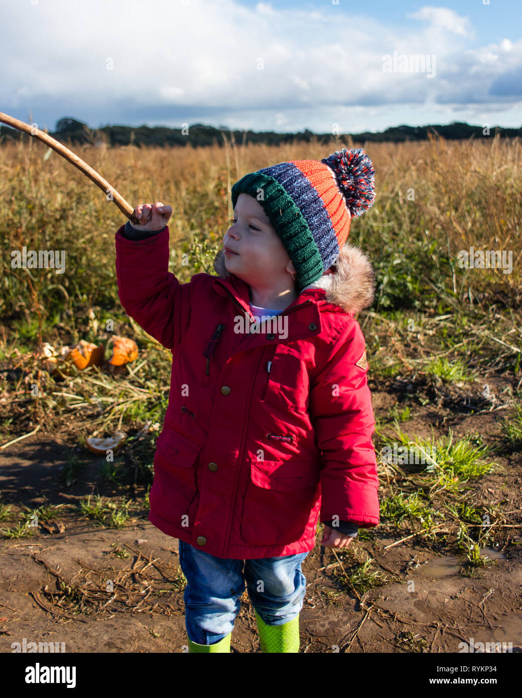 Zwei Jahre alten Spielen mit einem Stock in die Landschaft trägt einen roten Mantel an Bobble hat Stockfoto