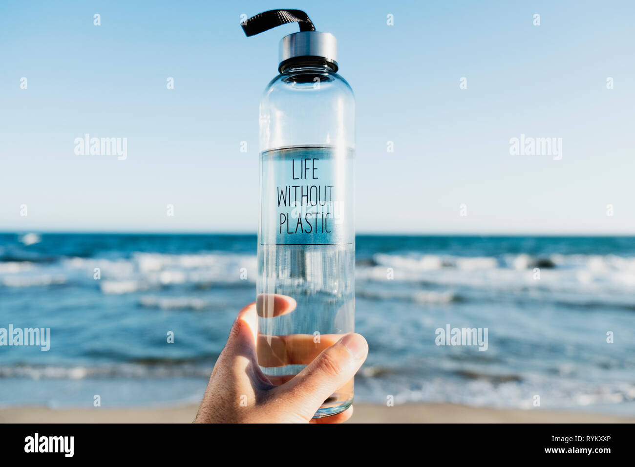 Nahaufnahme von einem kaukasischen Mann hält ein Glas wiederverwendbare Trinkflasche mit dem Text leben ohne Kunststoff in Es geschrieben, am Strand, mit dem Meer in der Stockfoto