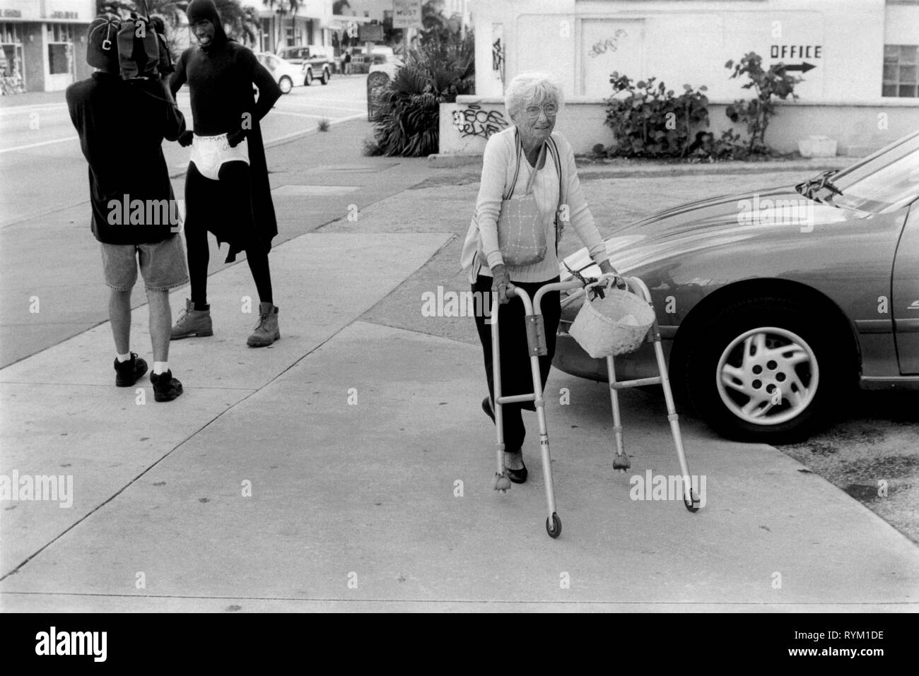 South Beach Miami, Florida USA 1999. Alte Frau mit Gehhilfe zu Fuß vorbei an einem afro-amerikanischen Mann, der wie ein super hero gekleidet ist. Er trägt seine unter Shorts über seine schwarzen Kostüm während gefilmt zu werden. 1990 s UNS HOMER SYKES. Stockfoto