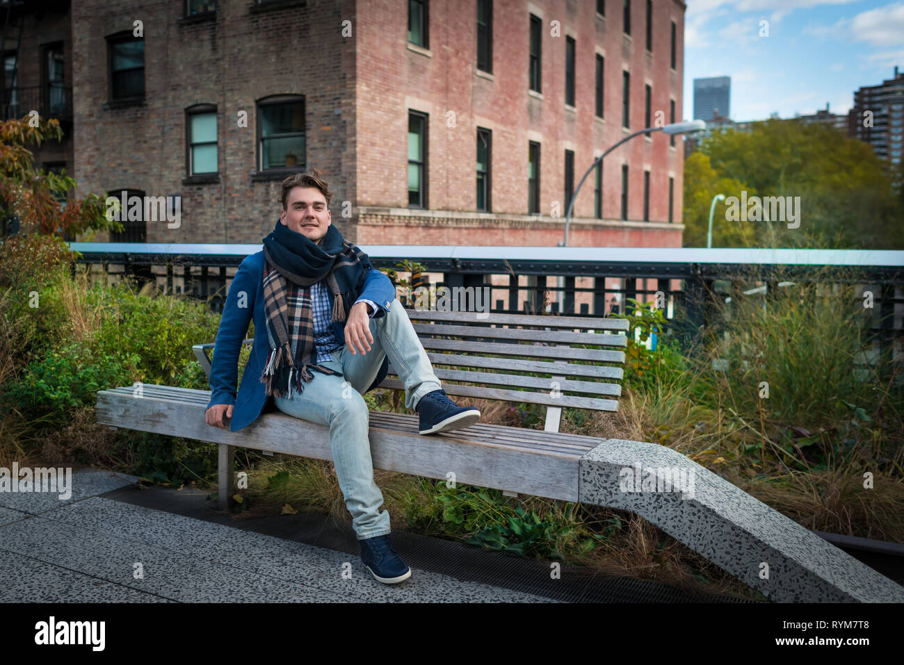 Junge eleganten Mann entspannen mit Blick auf Gebäude und Bäume in der High Line Park in New York City, USA. Hübscher Kerl sitzt auf der Bank und Suchen in der Kamera. Stockfoto