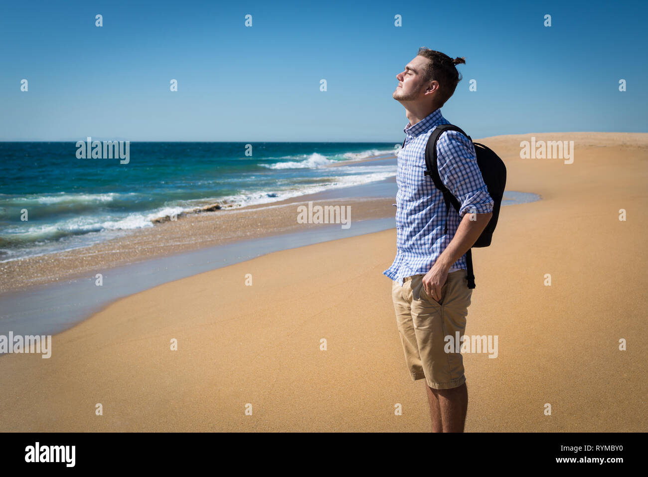 Glückliche junge Mann im blauen Hemd mit Rucksack mit sonnigen Sommertag am Strand in Kalifornien, USA. Hübscher Kerl alleine stehen mit Blick auf den Ozean Stockfoto