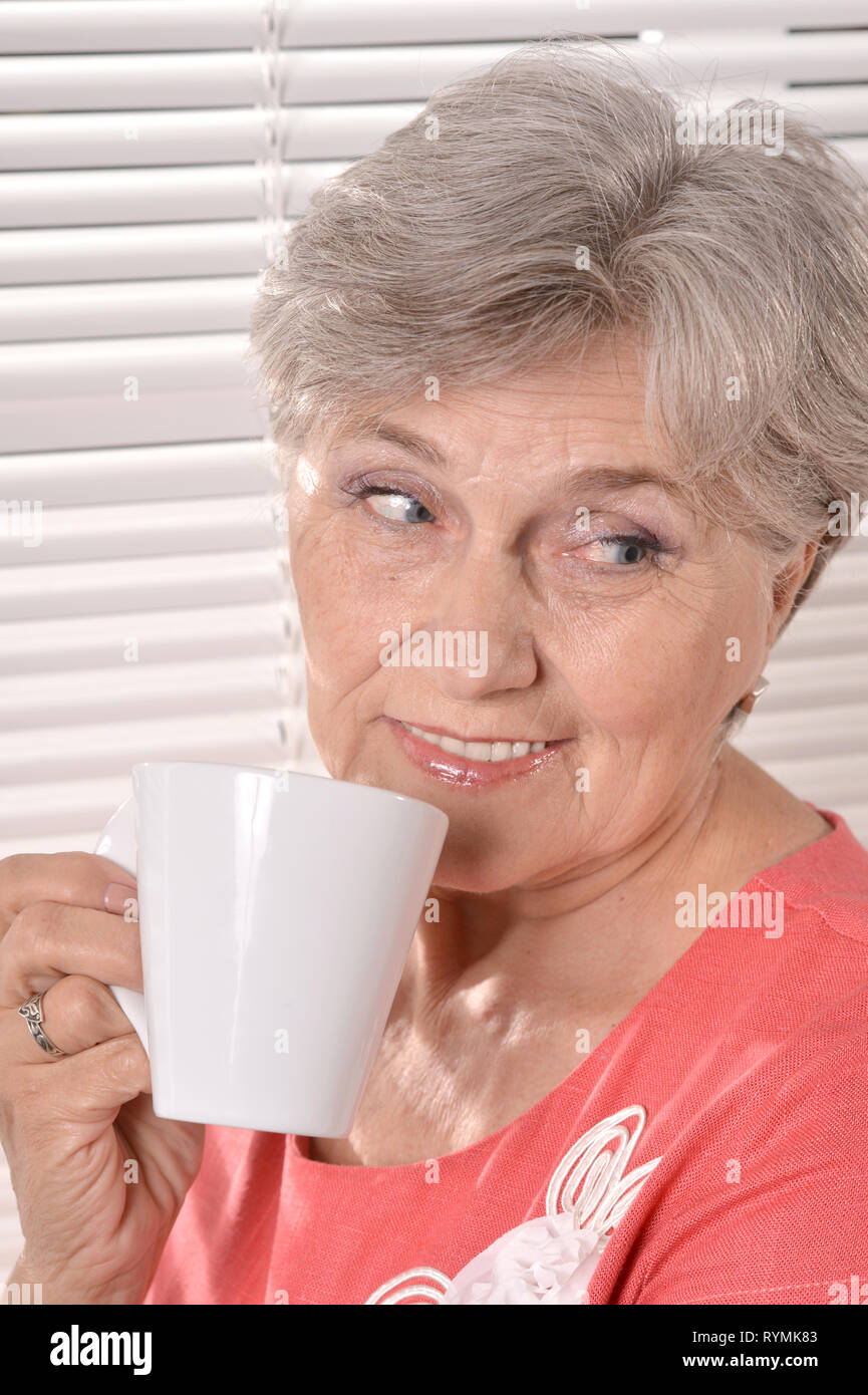 Gerne ältere Frau trinkt Tee auf weißen Jalousien Hintergrund Stockfoto