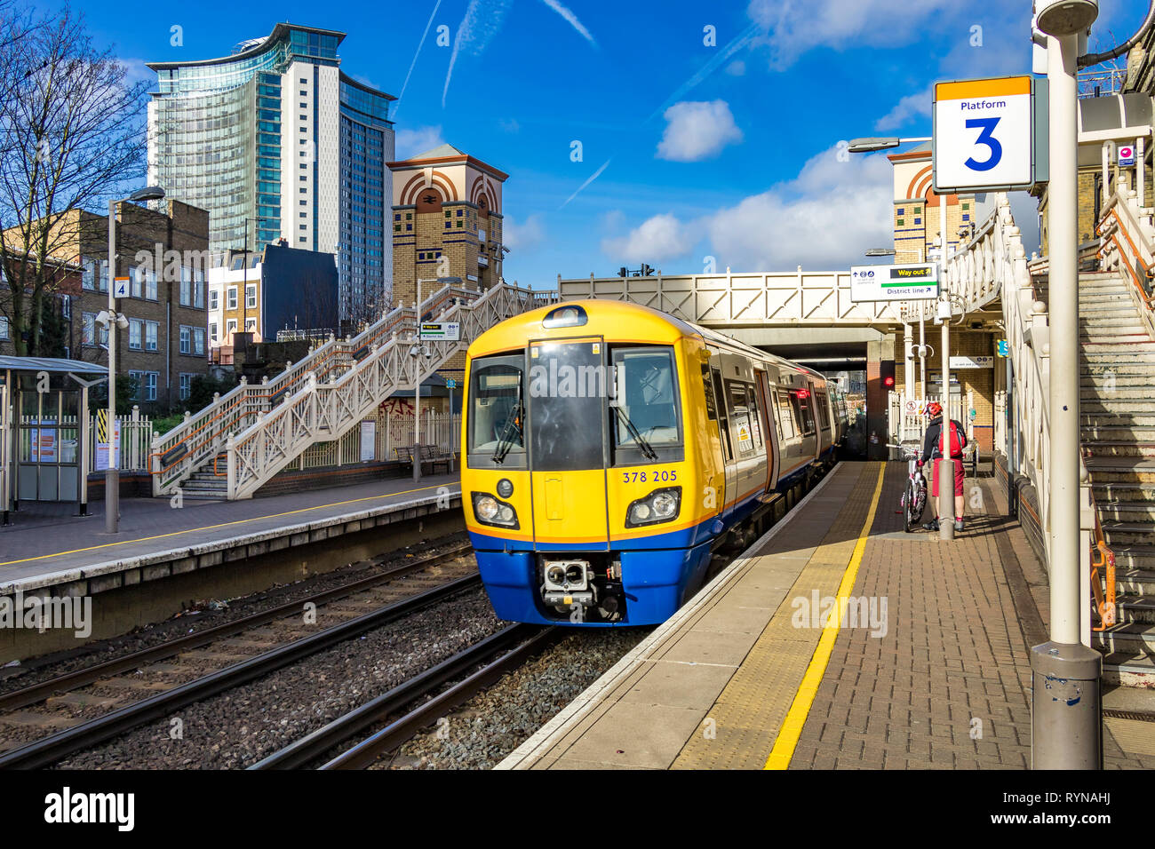 Ein London Overground Zug Richtung Clapham Junction fährt zum Bahnhof West Brompton mit dem Empress State Building in der Ferne, London, Großbritannien Stockfoto