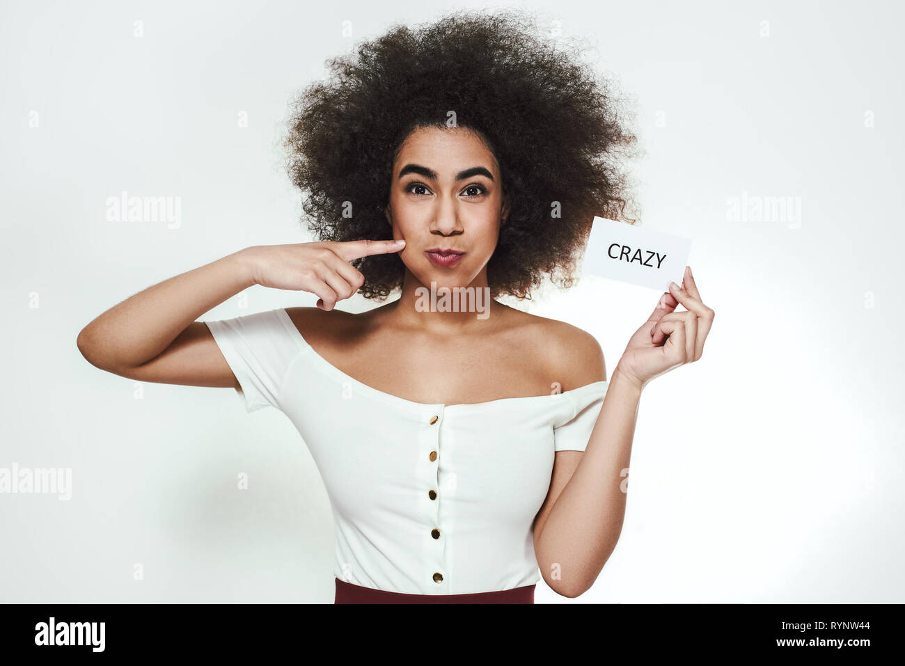 Spielerische. Lustig junge afrikanische Frau ihre Wange zu berühren und halten Sie ein Stück Papier ein, die sagen 'Crazy' ist, während gegen grauen Hintergrund stehen. Spaß-Konzept. Positive Emotionen Stockfoto
