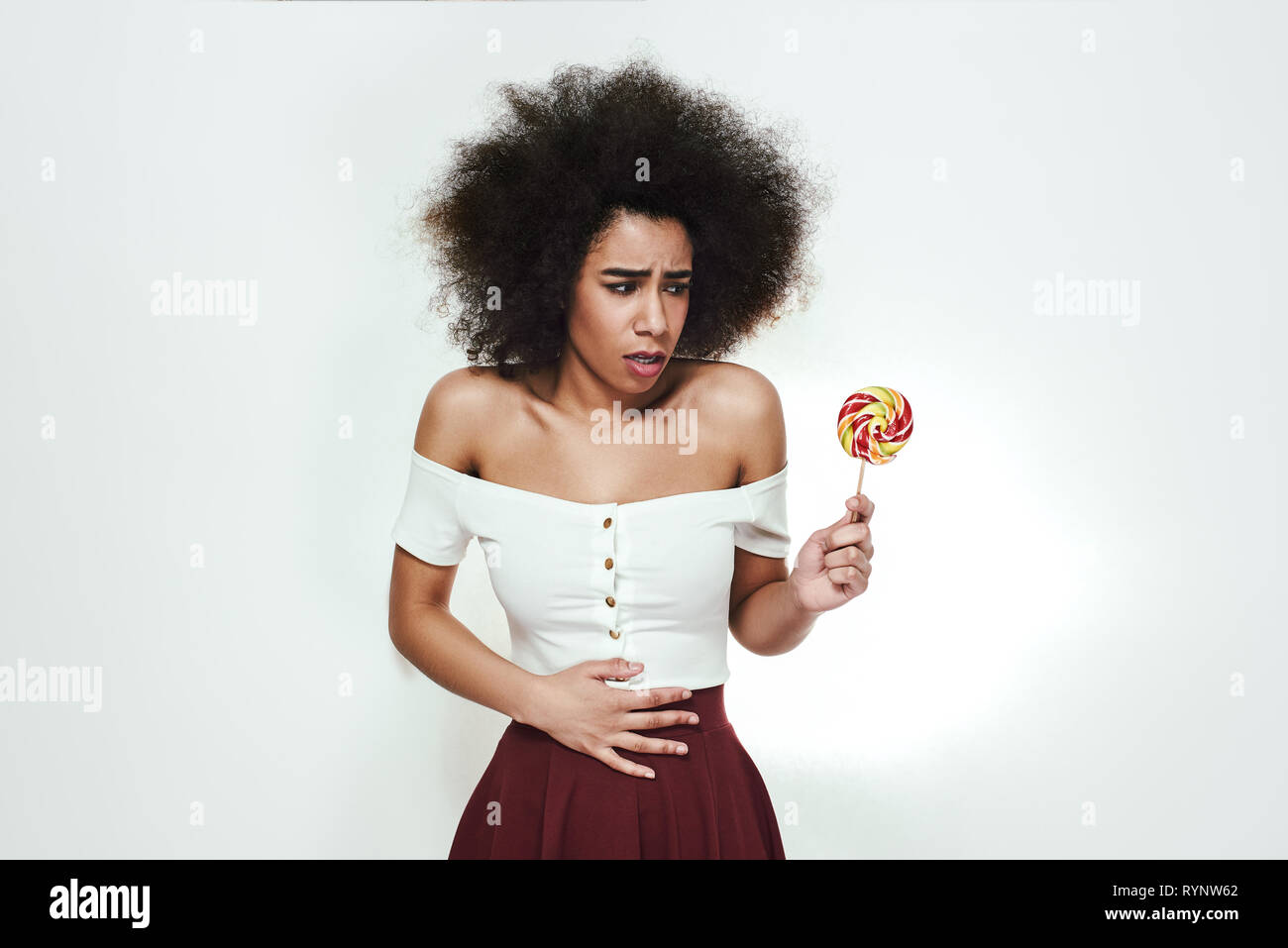 Nicht zu viel Süßigkeiten essen! Junge afro-amerikanische Frau mit bunten Lutscher in der Hand Bauchschmerzen, weil sie zu viel Süßigkeiten gegessen. Ungesunde Lebensmittel. Krankheit Konzept Stockfoto